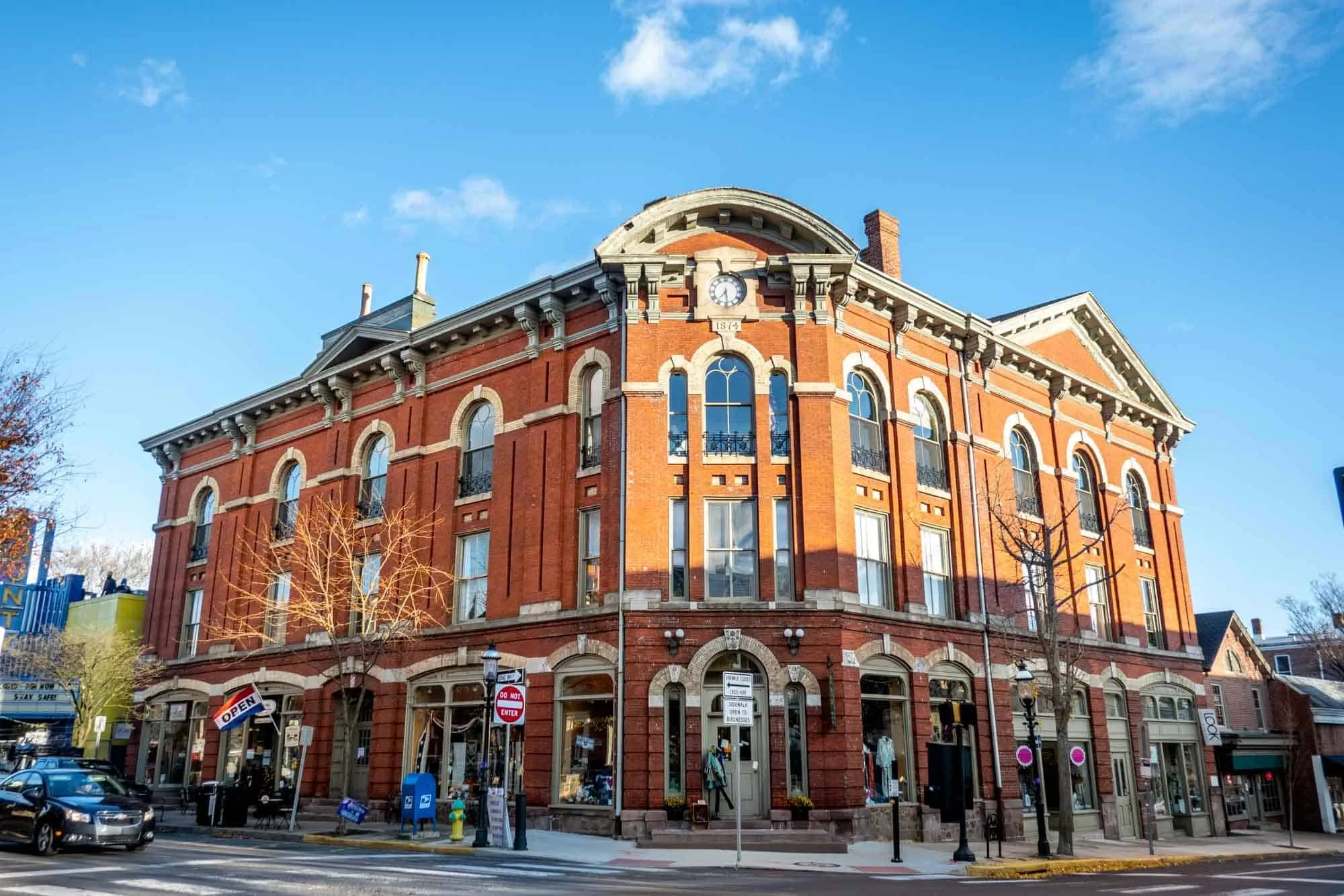 Red brick historic building with arched windows and clock on top, situated on a street corner with storefronts at street level and storefront signs, leafless trees, cars parked, and a clear blue sky.
