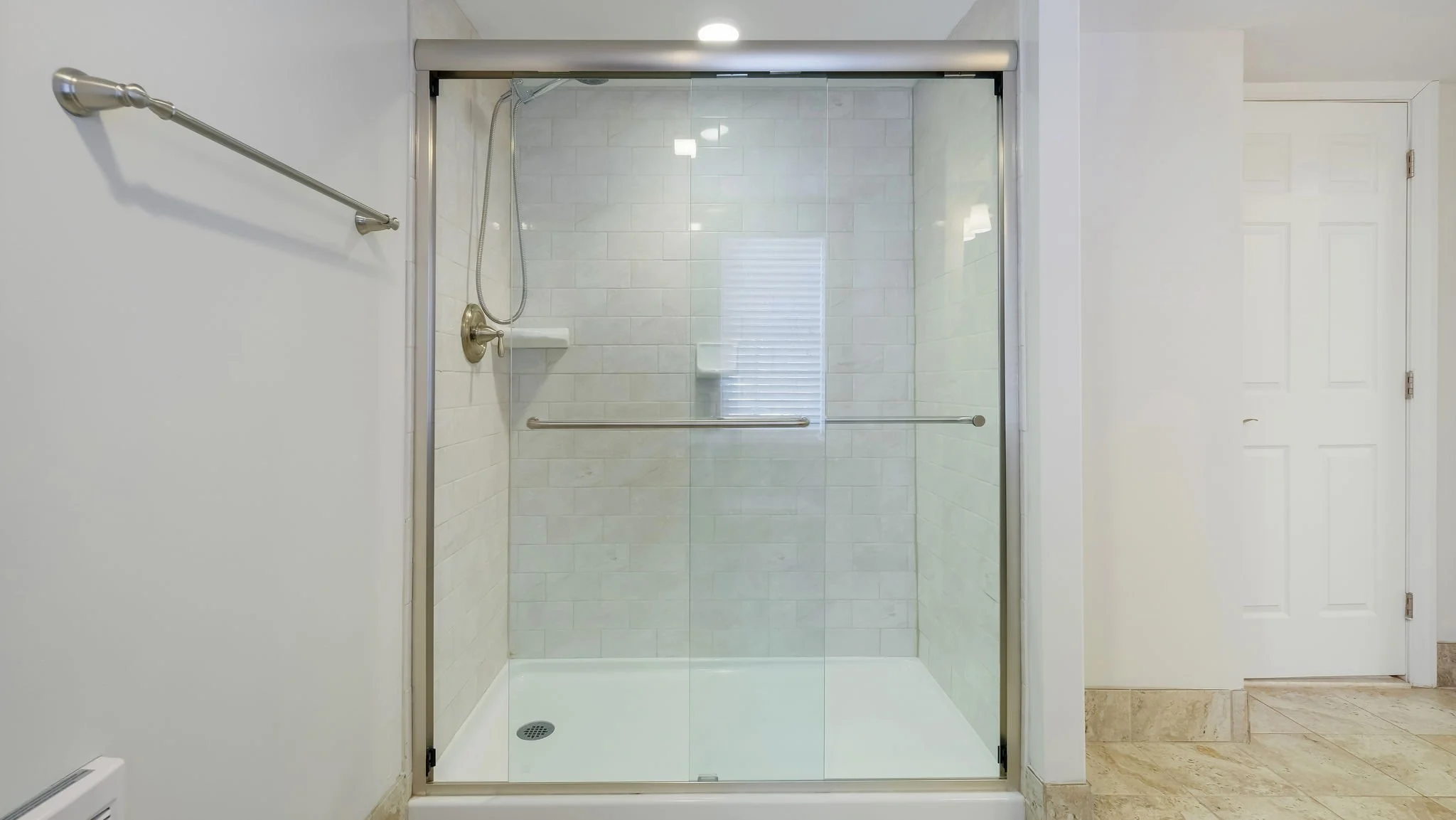 A glass shower enclosure with a sliding door in a bathroom, with beige tiled walls and a white shower tray, a showerhead, and a single shelf.