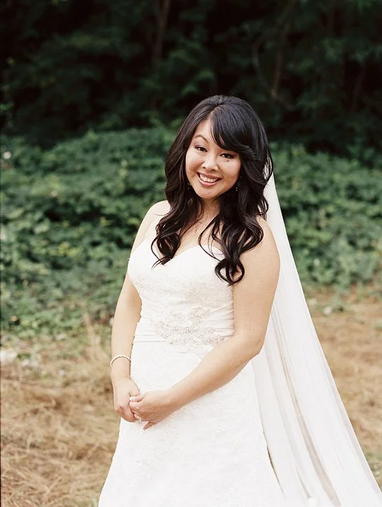 A woman in a white wedding dress smiling outdoors with trees and greenery in the background.