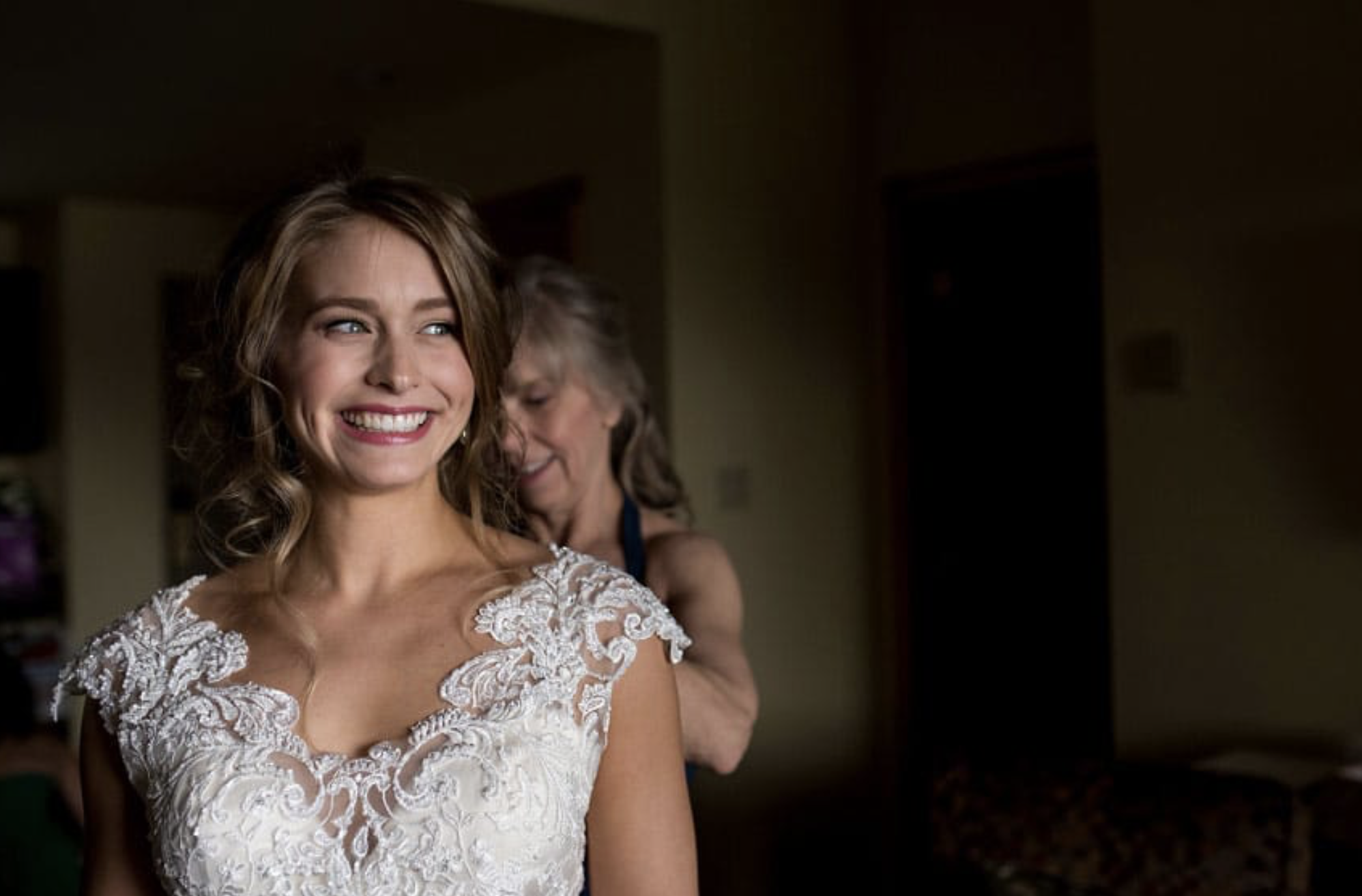 A young woman in a wedding dress smiling brightly as an older woman adjusts her gown in a warmly lit room.