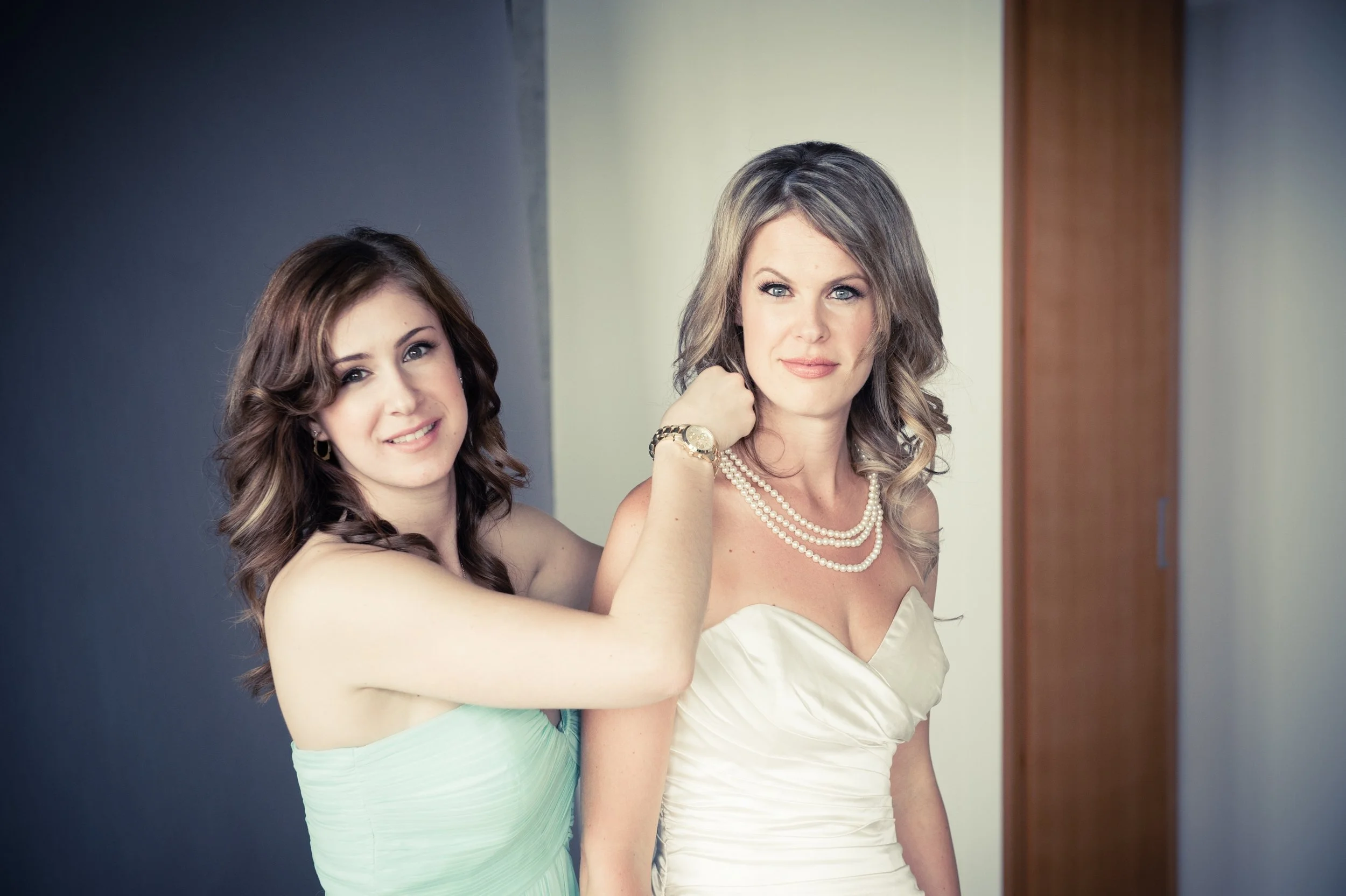 Two women in formal attire, one adjusting the other's pearl necklace, standing indoors against a neutral background.