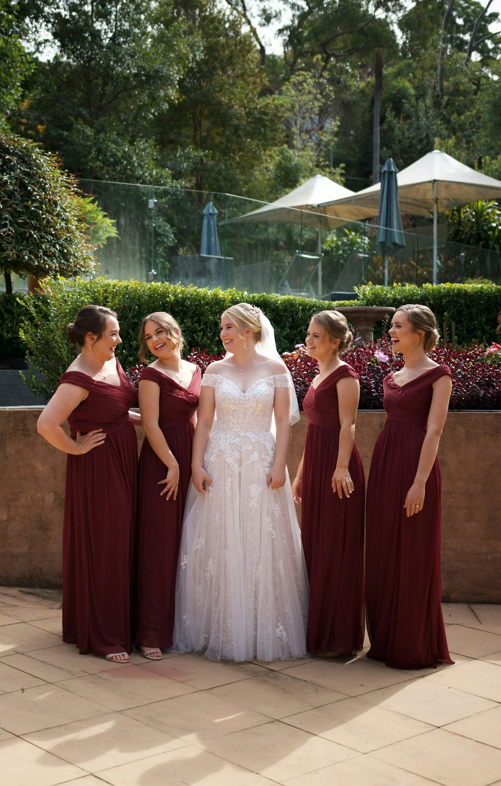 Bride in a white wedding dress standing with four bridesmaids in maroon dresses outdoors.