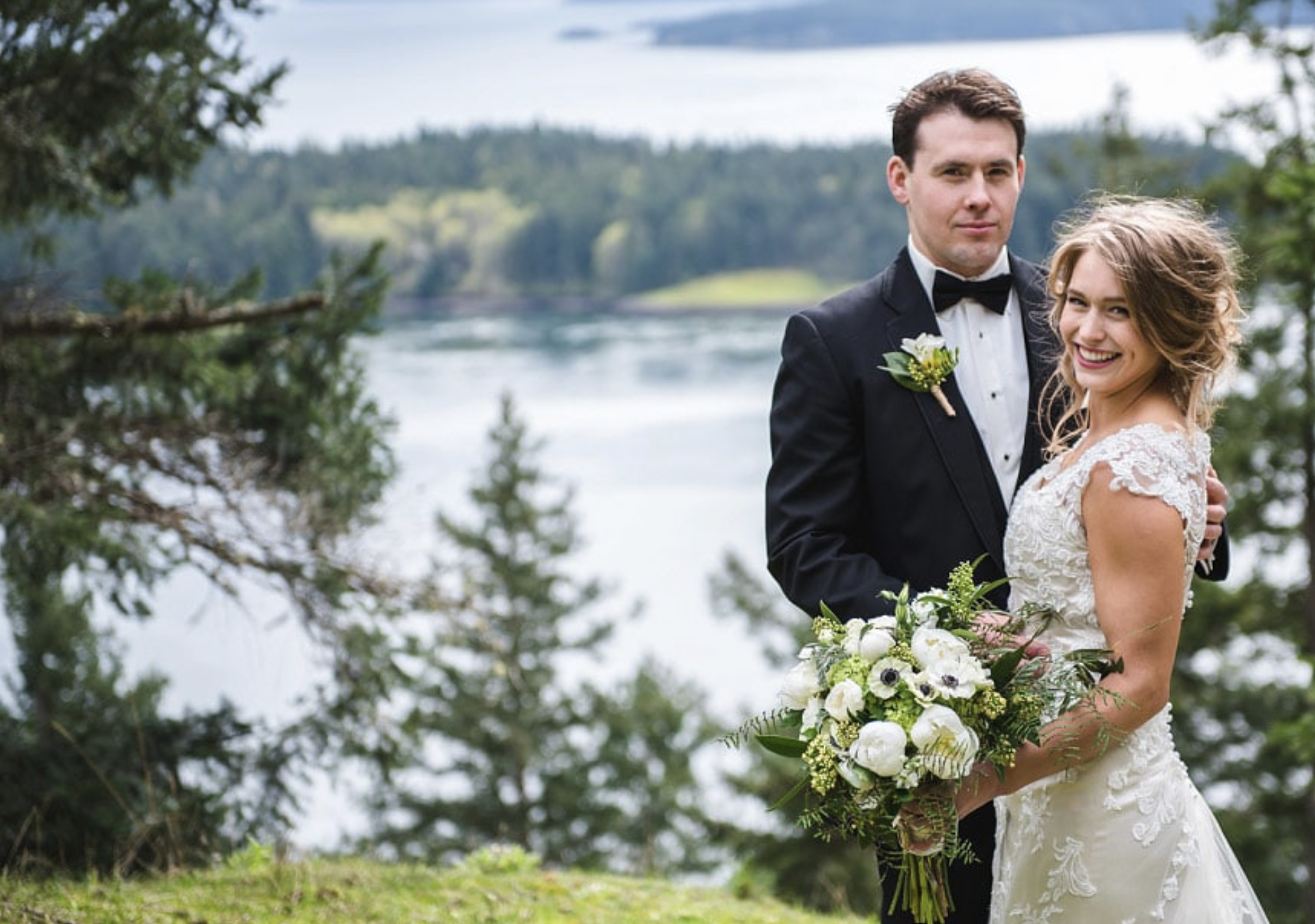 A newlywed couple standing outdoors near a lake with trees and hills in the background. The groom is in a black tuxedo with a boutonniere, and the bride is in a white lace wedding dress holding a bouquet of white and green flowers. The bride is smili