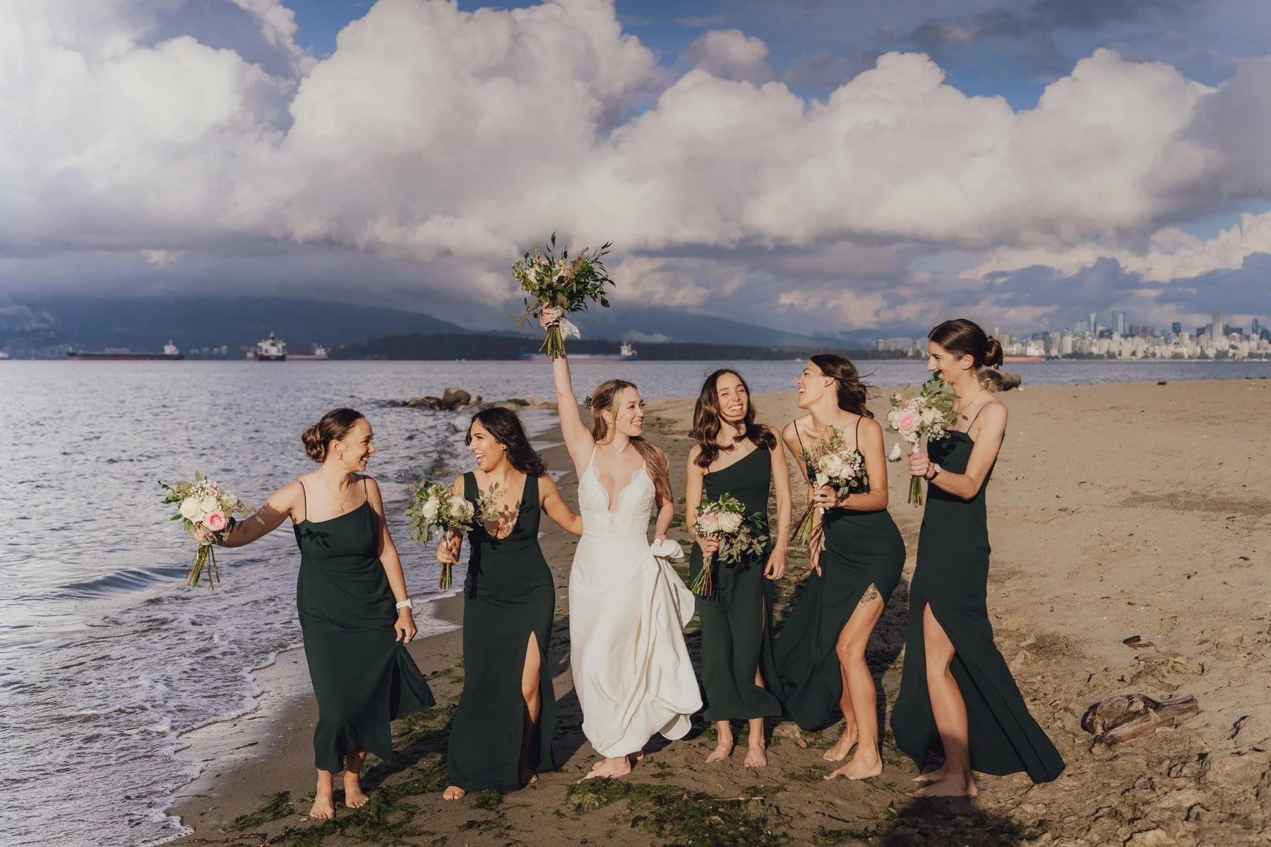 A bride and five bridesmaids walking on a beach, with the bride holding a bouquet raised in the air and smiling, while the bridesmaids hold bouquets and smile. The scene is set against a backdrop of water, ships, distant mountains, and a city skyline