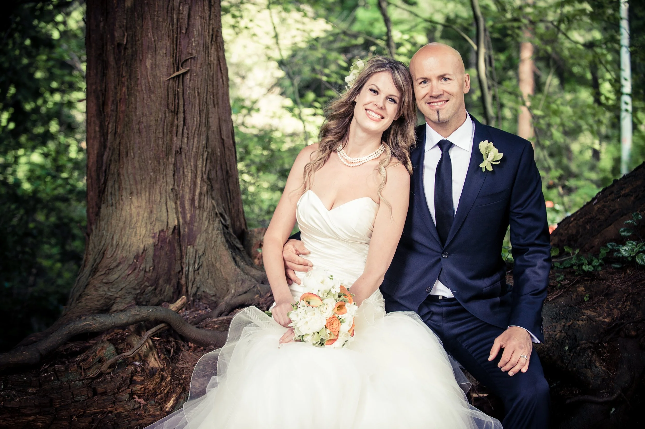 A bride and groom sitting on a fallen tree trunk in a forest, smiling and posing for a wedding photo. The bride is wearing a strapless white wedding gown, pearl necklace, and holding a bouquet of white and orange flowers. The groom is wearing a navy 