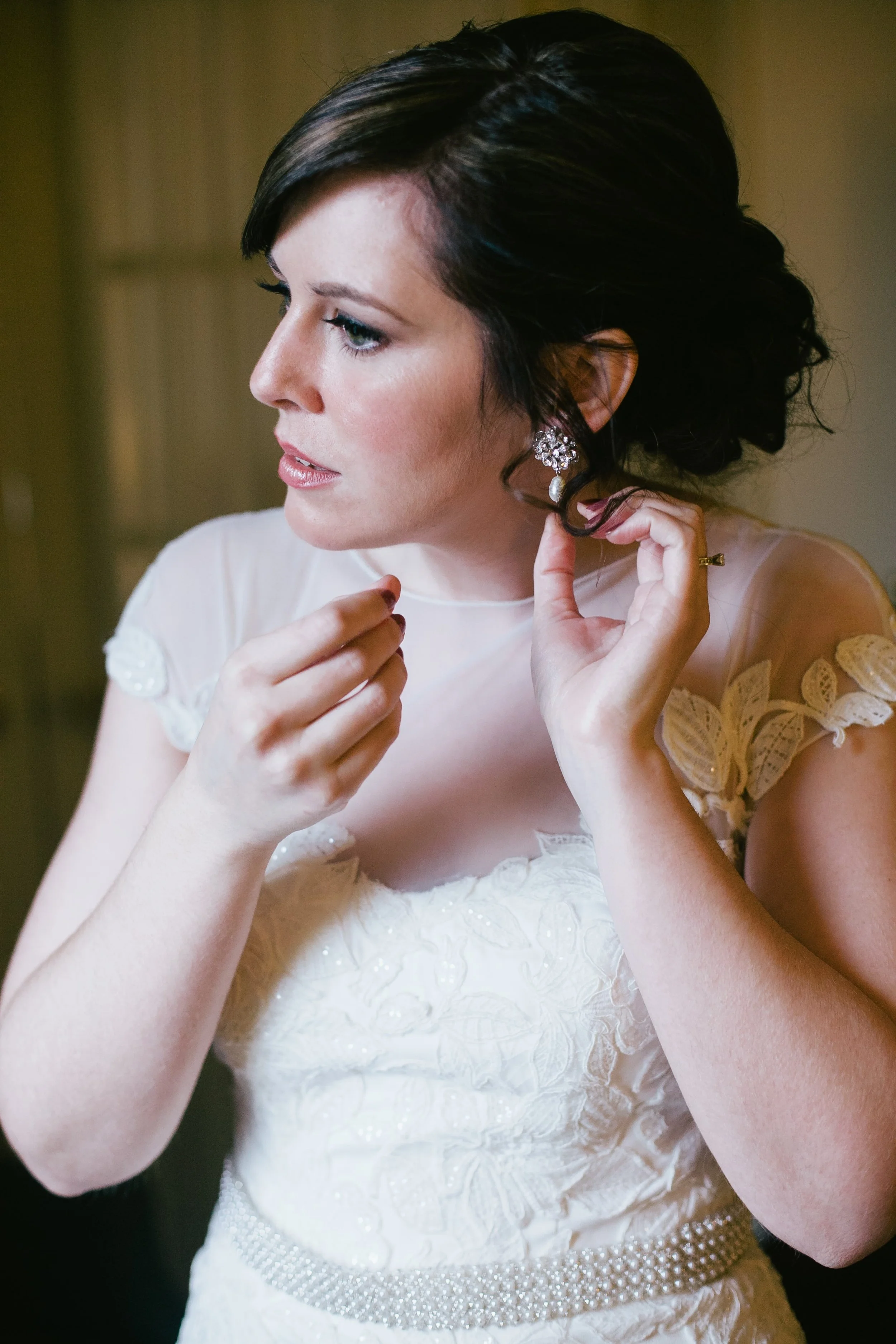 A woman with short dark hair dressing up in a white lace dress, putting on earrings.