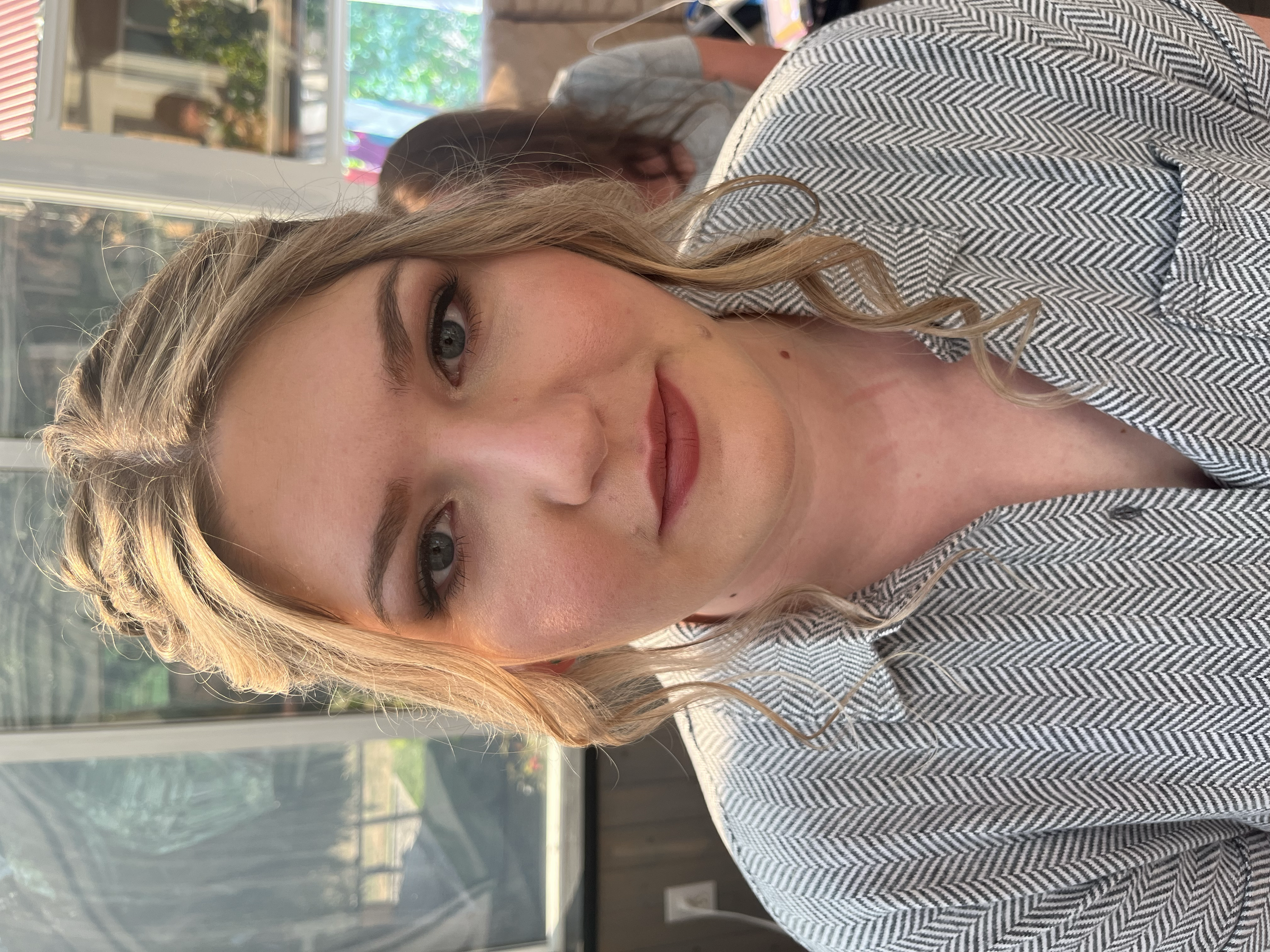 Close-up of a woman with wavy blonde hair, blue eyes, wearing makeup, and a gray patterned shirt, sitting indoors near large windows with a view of a patio.