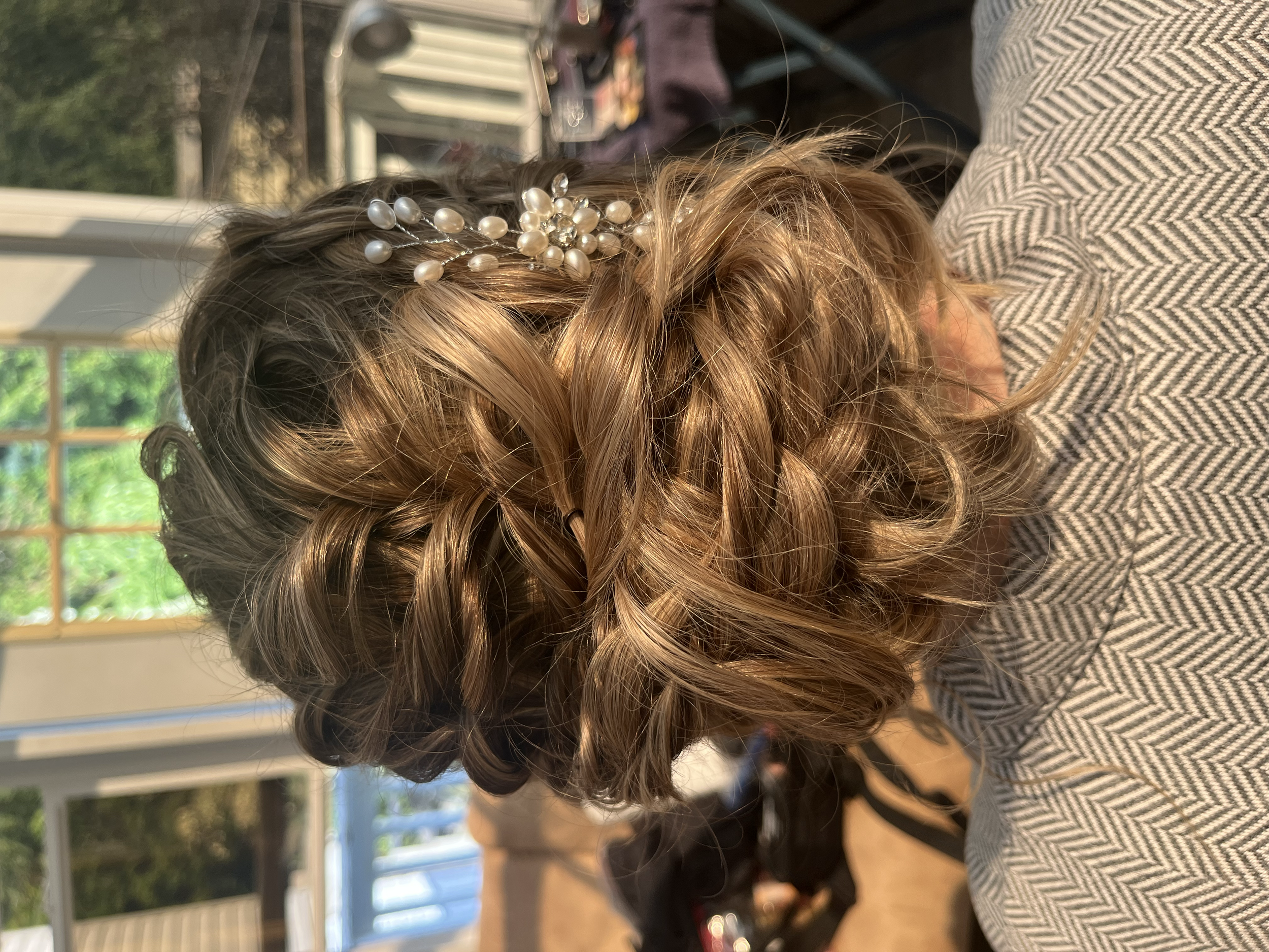 Back view of a woman with styled, curly blonde hair decorated with a pearl and rhinestone hair accessory, wearing a herringbone patterned garment, indoors near a window with sunlight