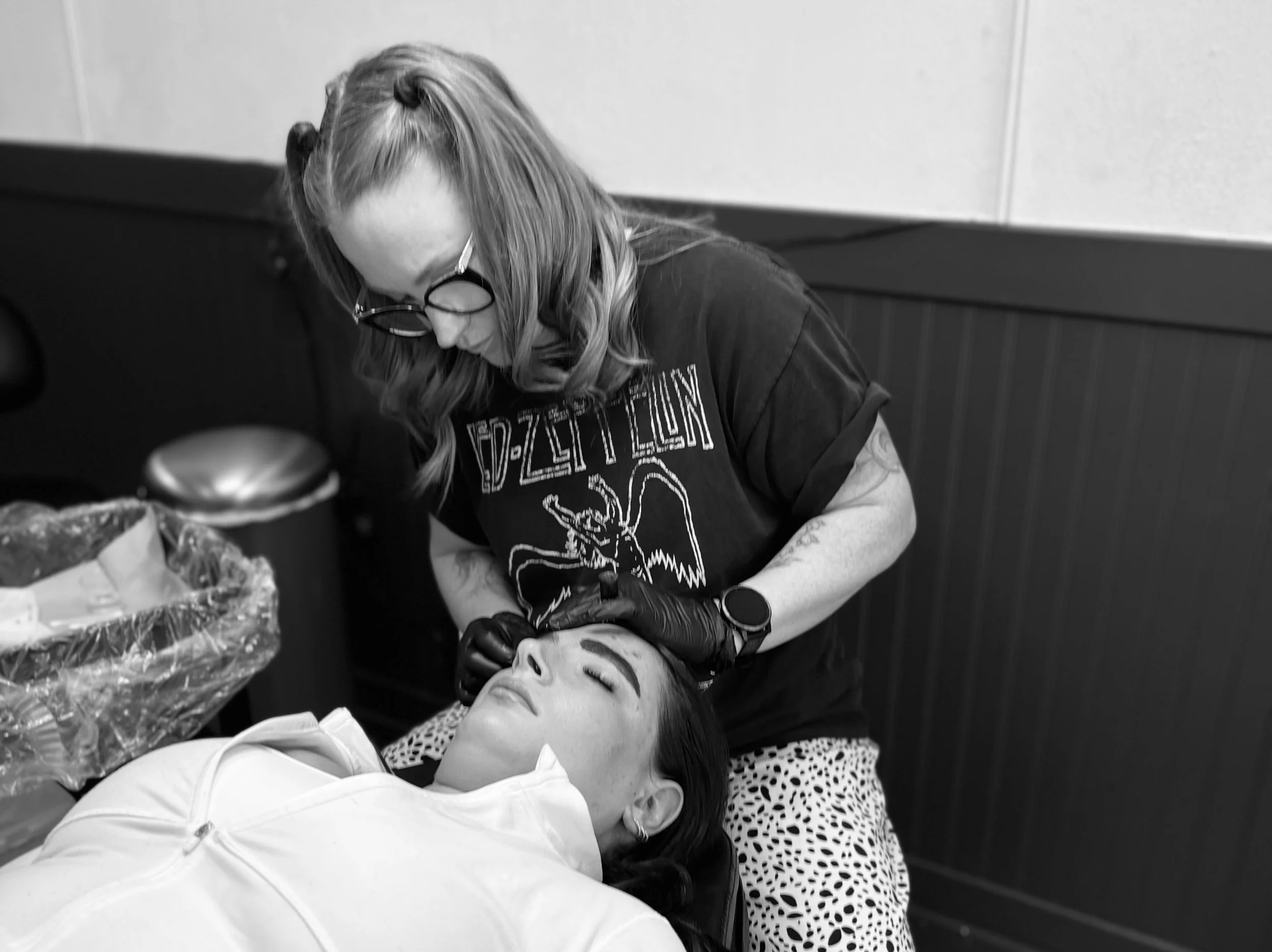 A woman is lying down with eyes closed, while another woman, wearing glasses and a Led Zeppelin t-shirt, is tattooing her face in a tattoo studio.