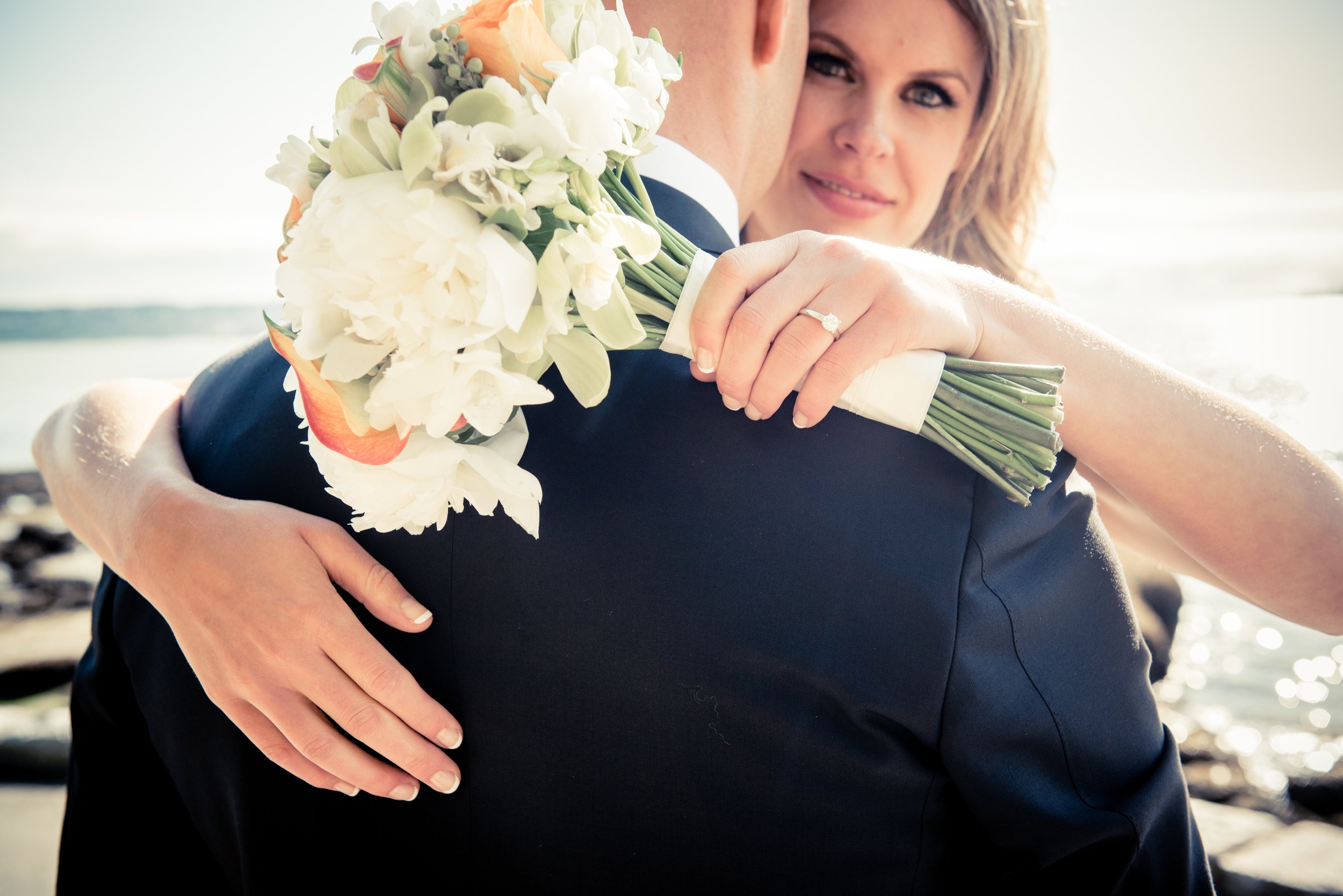 A bride and groom embrace on the beach, with the bride holding a bouquet of white and orange flowers, and showing her engagement ring. The bride's face is visible.