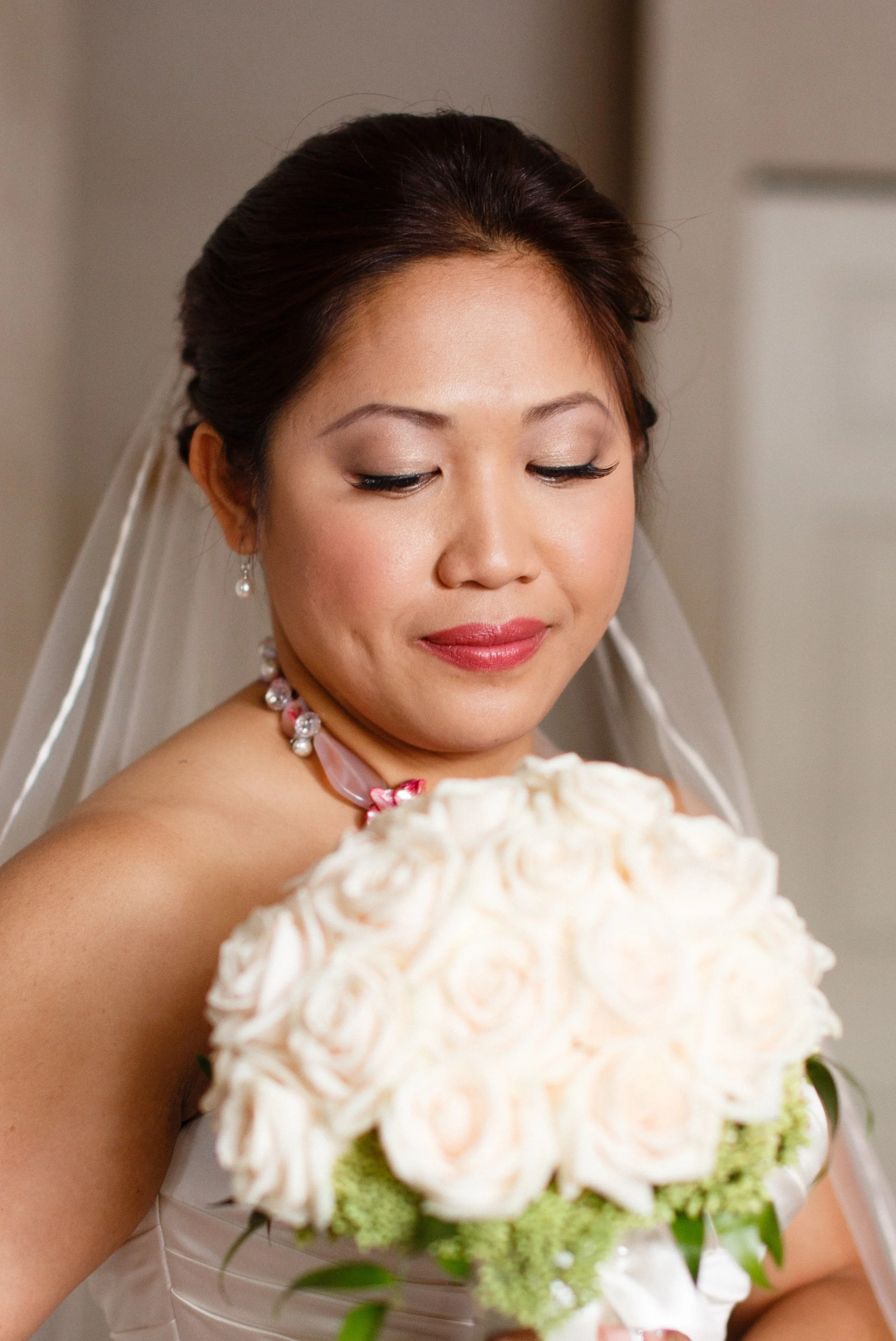 Close-up of a bride holding a bouquet of white roses on her chest, wearing a wedding dress, pearl necklace, earrings, and a veil, with her eyes closed and smiling softly.