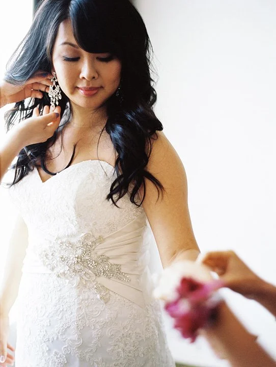 A bride in a white wedding dress with lace details and pearl embellishments, having her earrings adjusted by someone else.