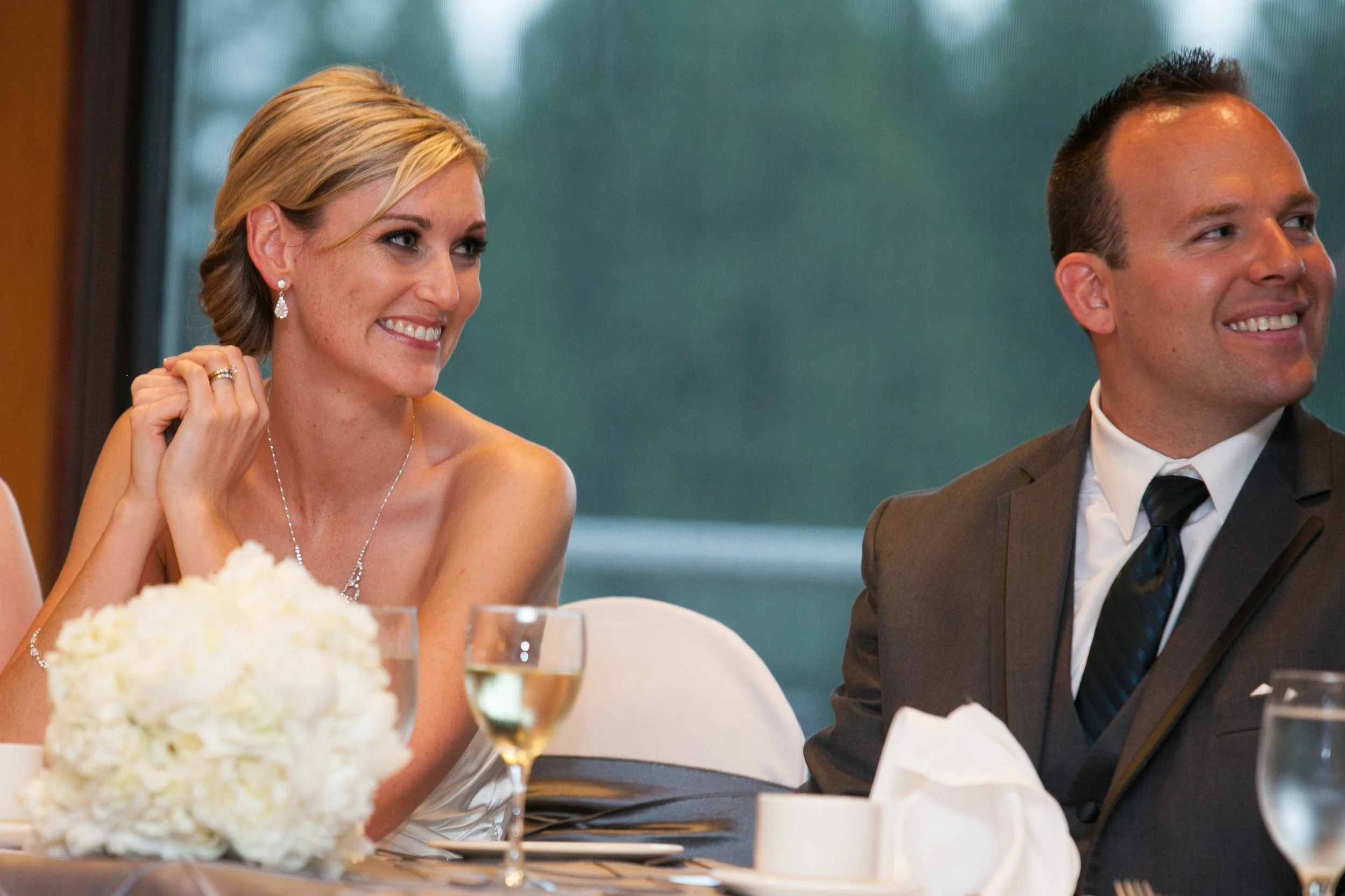 A woman with short blonde hair wearing earrings and a necklace, smiling and sitting at a table with a man in a dark suit and tie, in a restaurant or banquet setting.