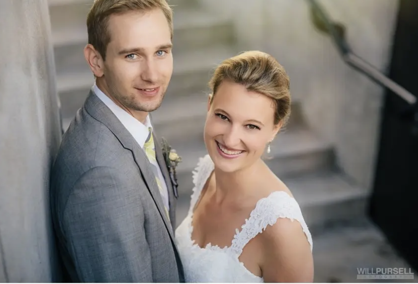 A smiling bride and groom standing close together on a staircase, dressed in wedding attire. The groom is wearing a gray suit with a light-colored shirt and a tie, while the bride is wearing a white lace wedding dress with puffed sleeves and pearl ea