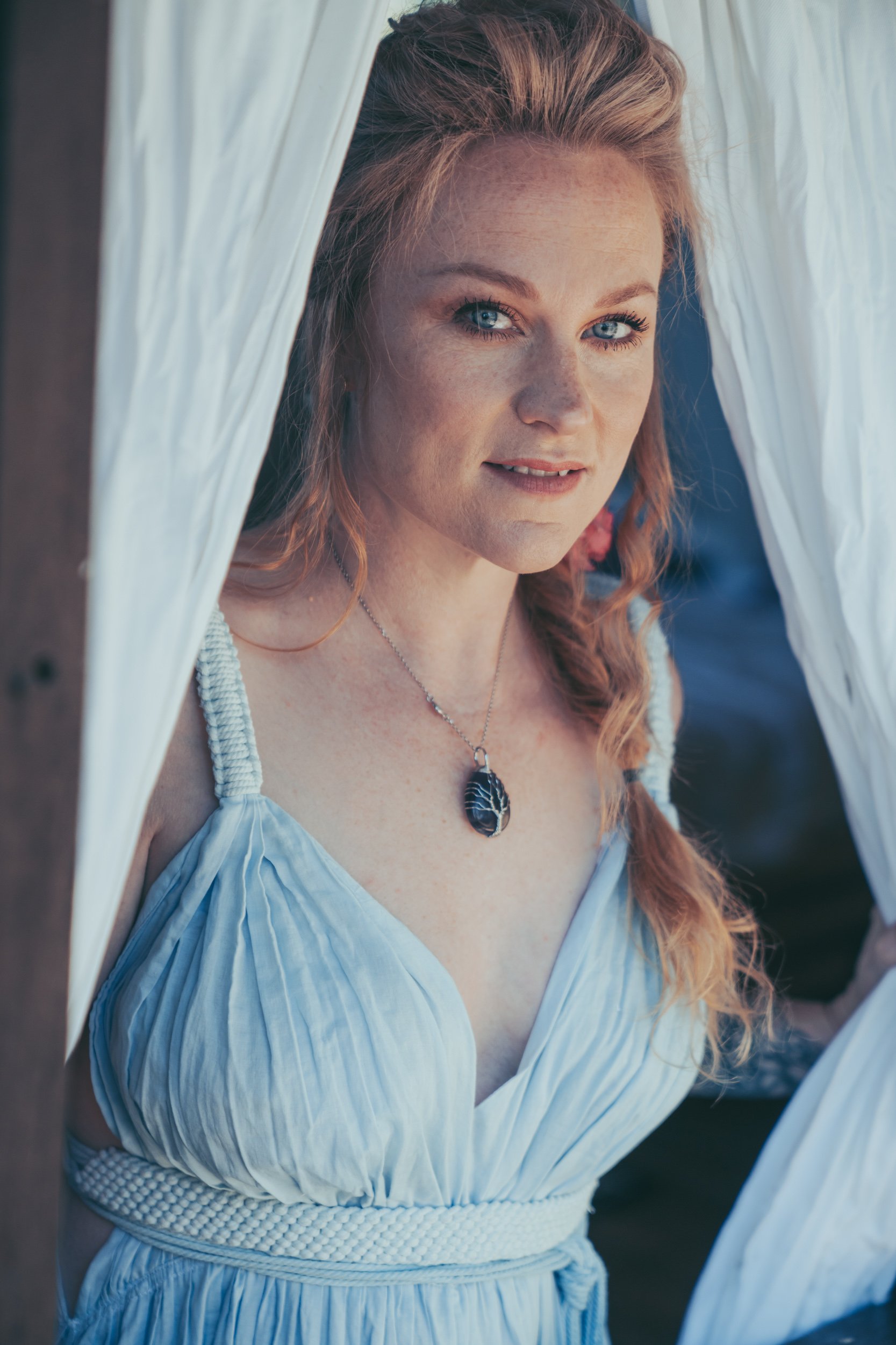 A woman with red hair and blue eyes looking out from behind white curtains, wearing a light blue dress and a black gemstone necklace.