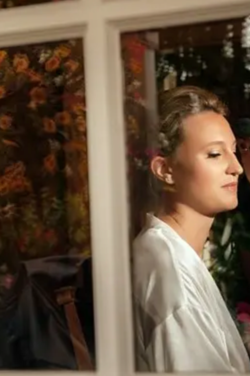 A young woman with braided hair looking out a window at night, with a floral background visible through the glass.