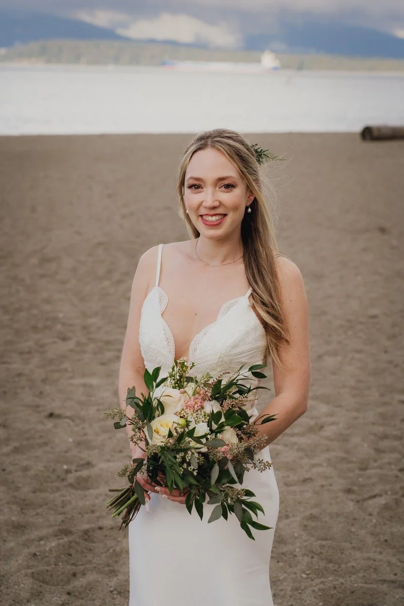 A smiling bride in a white wedding dress holding a bouquet of flowers on a beach with water and mountains in the background.