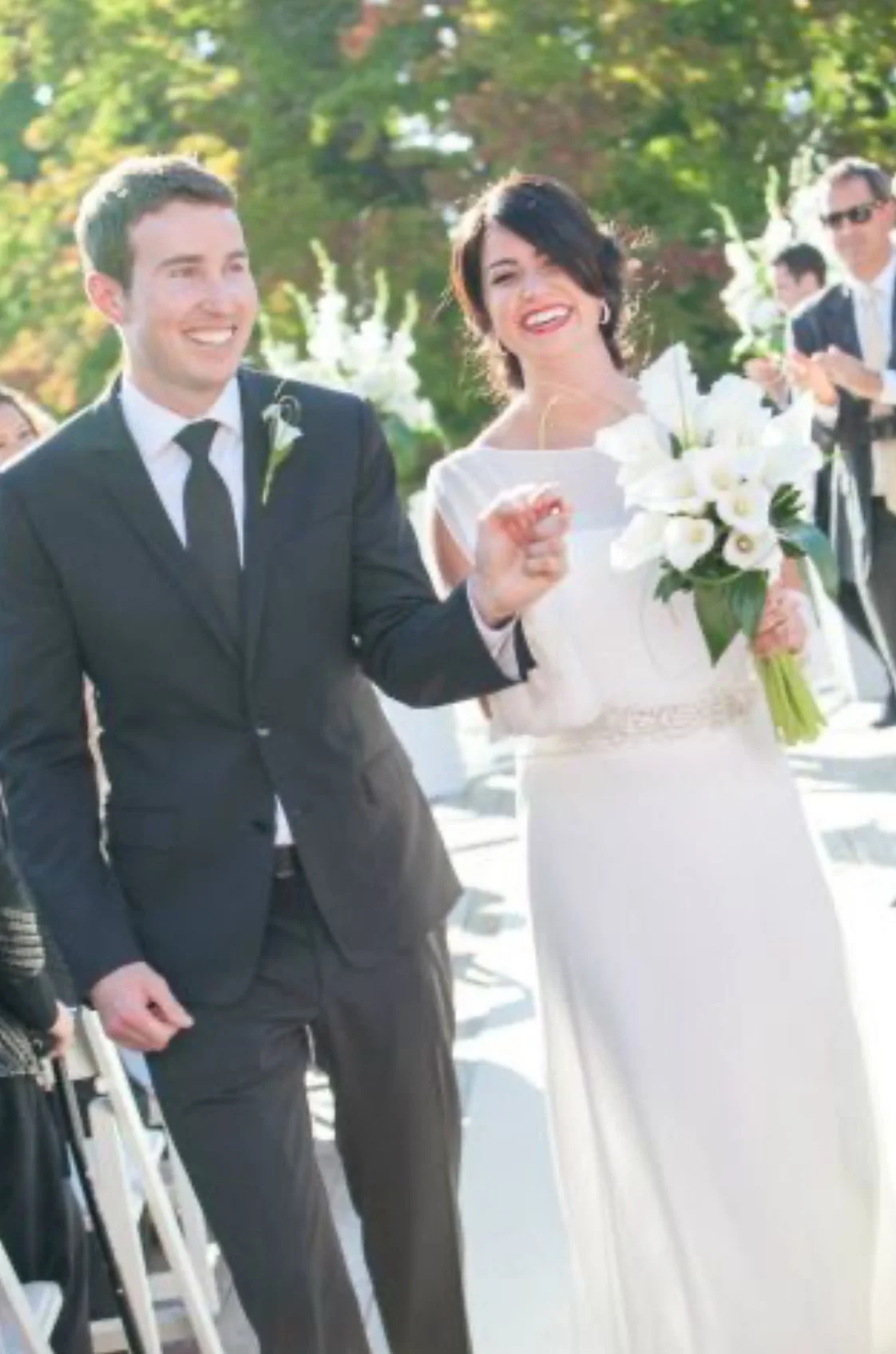 A smiling couple dressed in wedding attire walking outdoors, the man in a suit and the woman in a white dress holding a bouquet of flowers.
