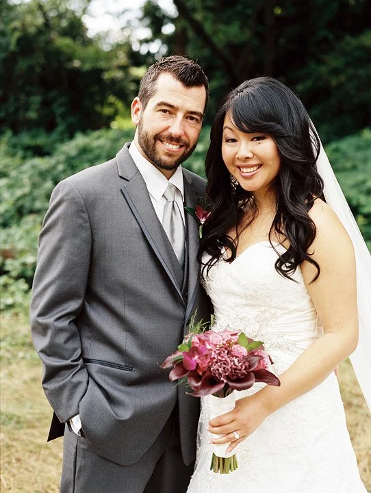 A newlywed couple standing outdoors with trees in the background, dressed in wedding attire, smiling and holding a bouquet of purple and pink flowers.