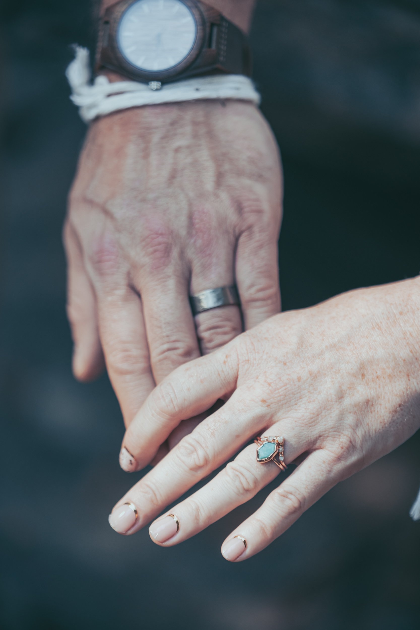 Close-up of two hands with wedding rings, one of which is wearing a watch and the other a ring on the finger with a gemstone. The hands are gently placed together.