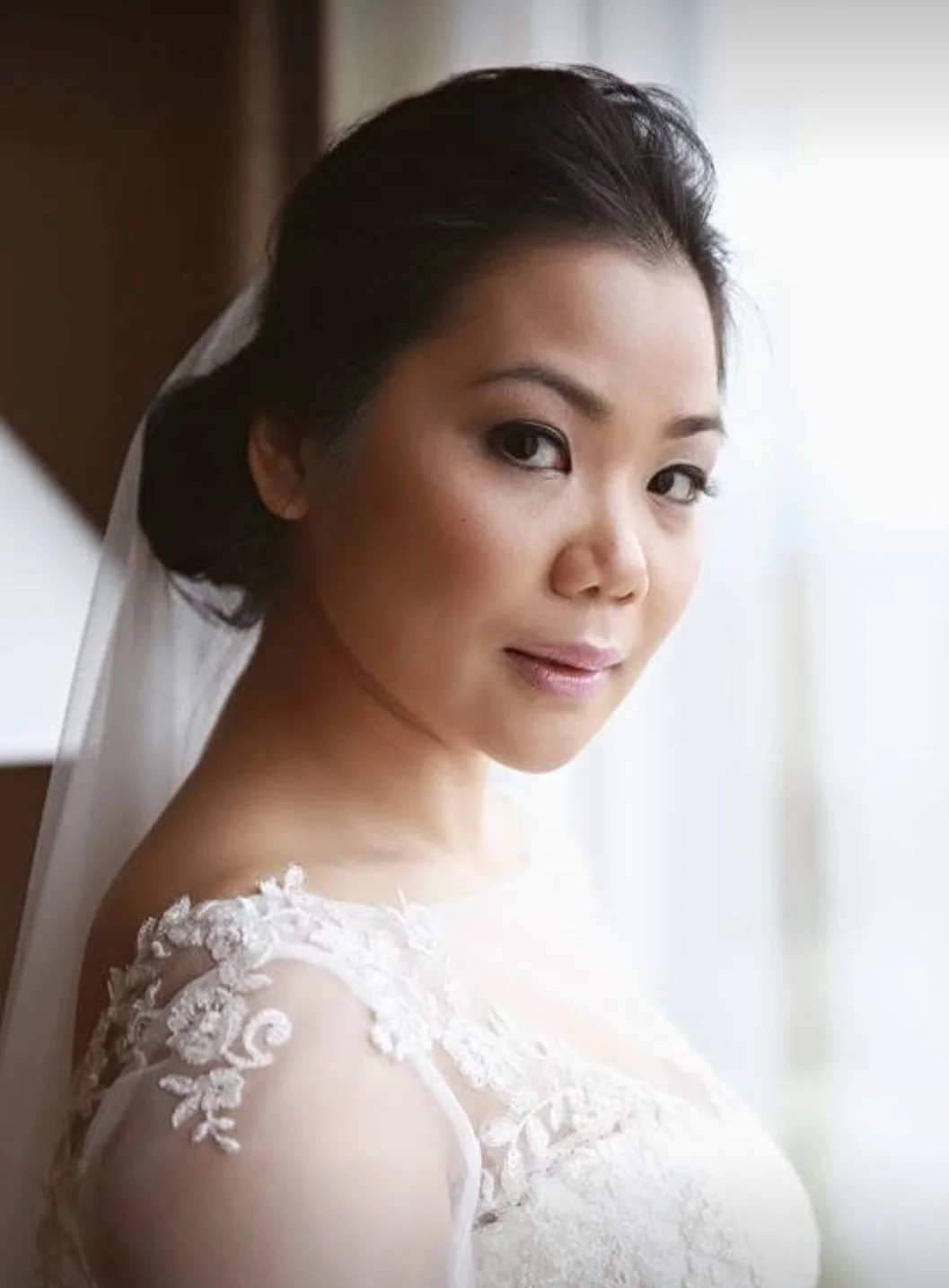 A bride with dark hair styled in an elegant updo, wearing a lace wedding dress and veil, looking over her shoulder.