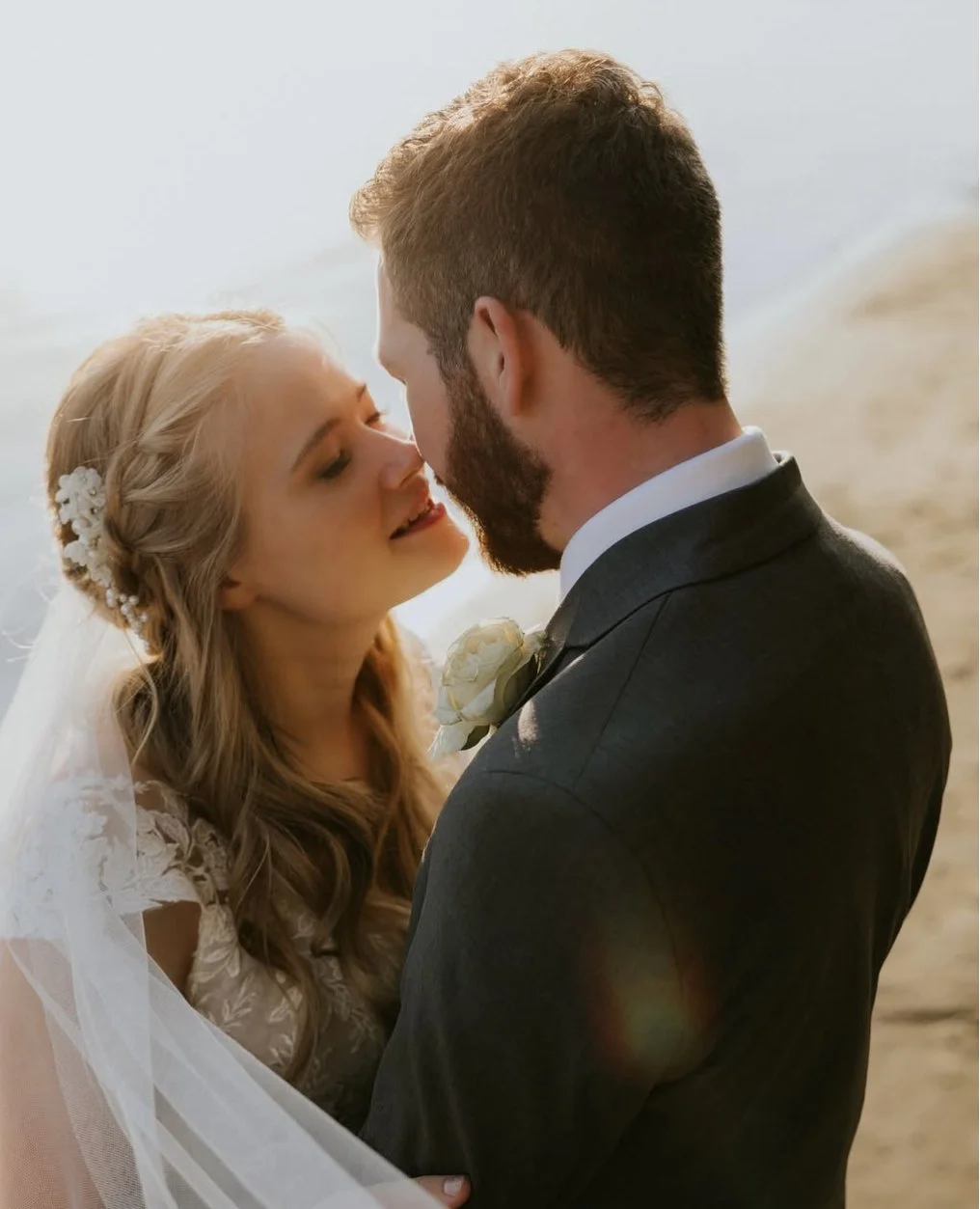 A bride and groom sharing a close moment on the beach, with the bride wearing a lace wedding dress and floral hair accessories, and the groom in a dark suit with a boutonniere, during sunset.