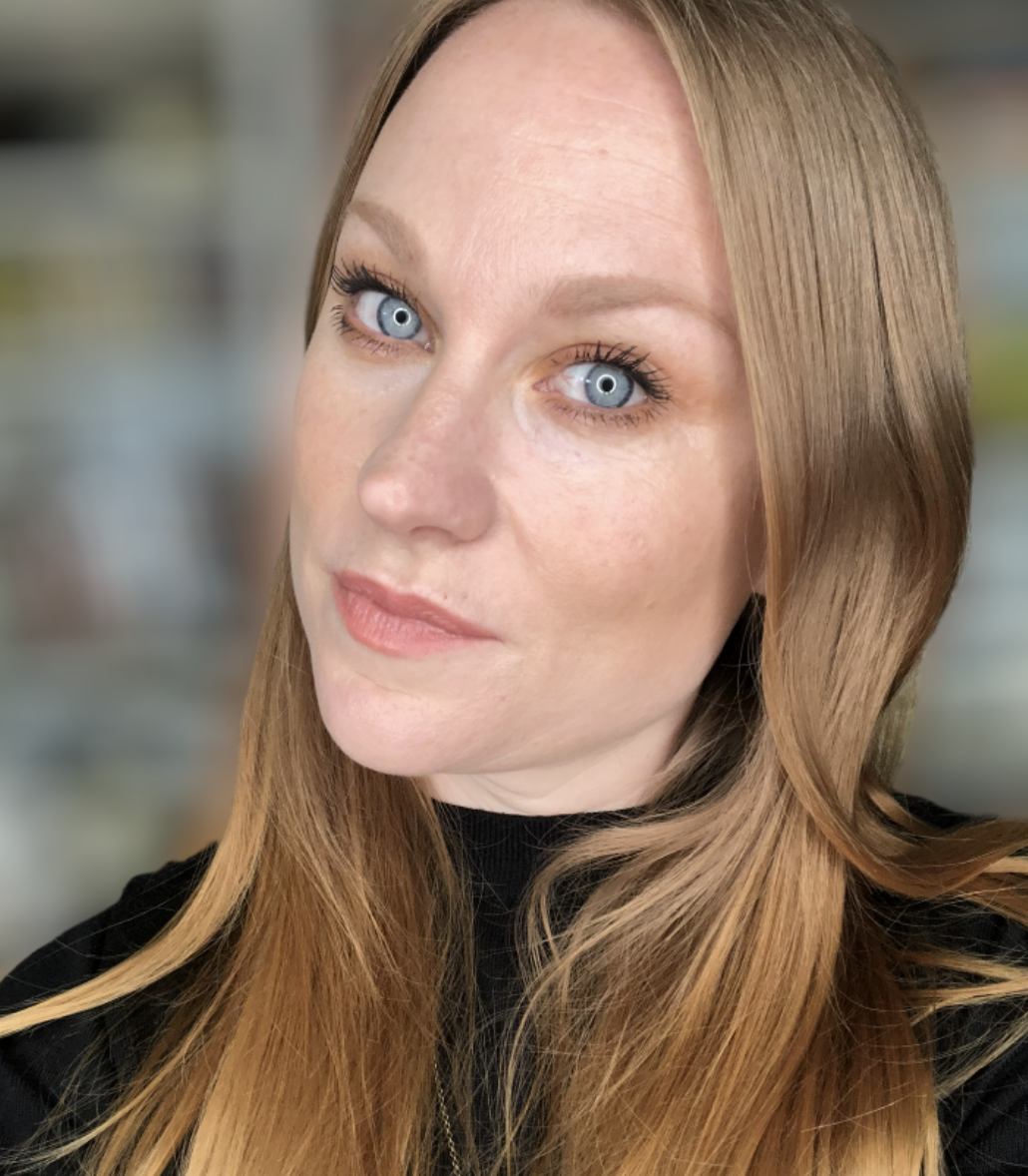 A woman with long ginger hair and blue eyes, wearing a black top, looking at the camera with a slight smile, indoors with a blurred background.
