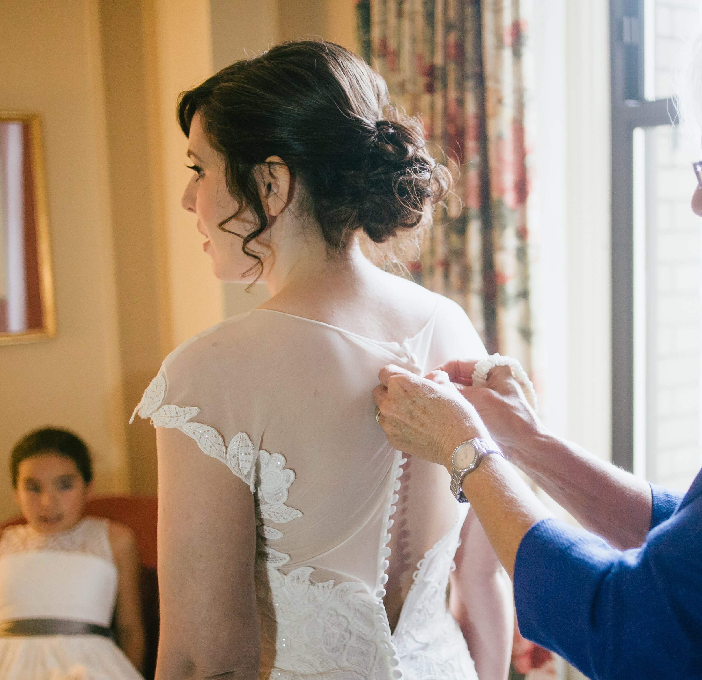 A woman in a wedding dress is having her dress buttoned by another person, with a young girl sitting in the background.