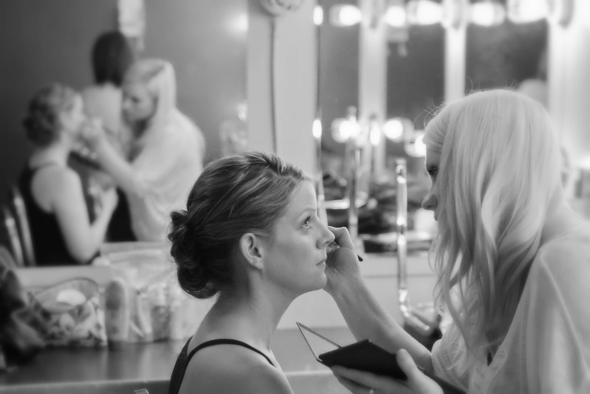 A woman applies makeup to another woman in a room with mirrors and lights, with a blurred background of two women talking.