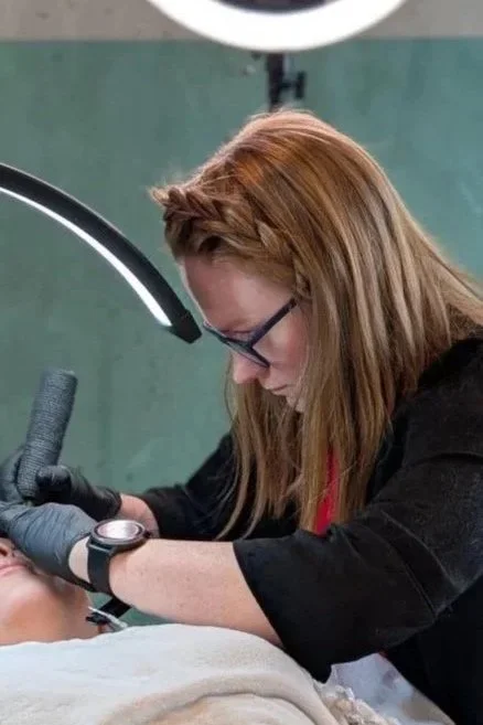 A woman with red hair and glasses performing a cosmetic or medical procedure on a patient's face, using professional tools and working under a ring light.