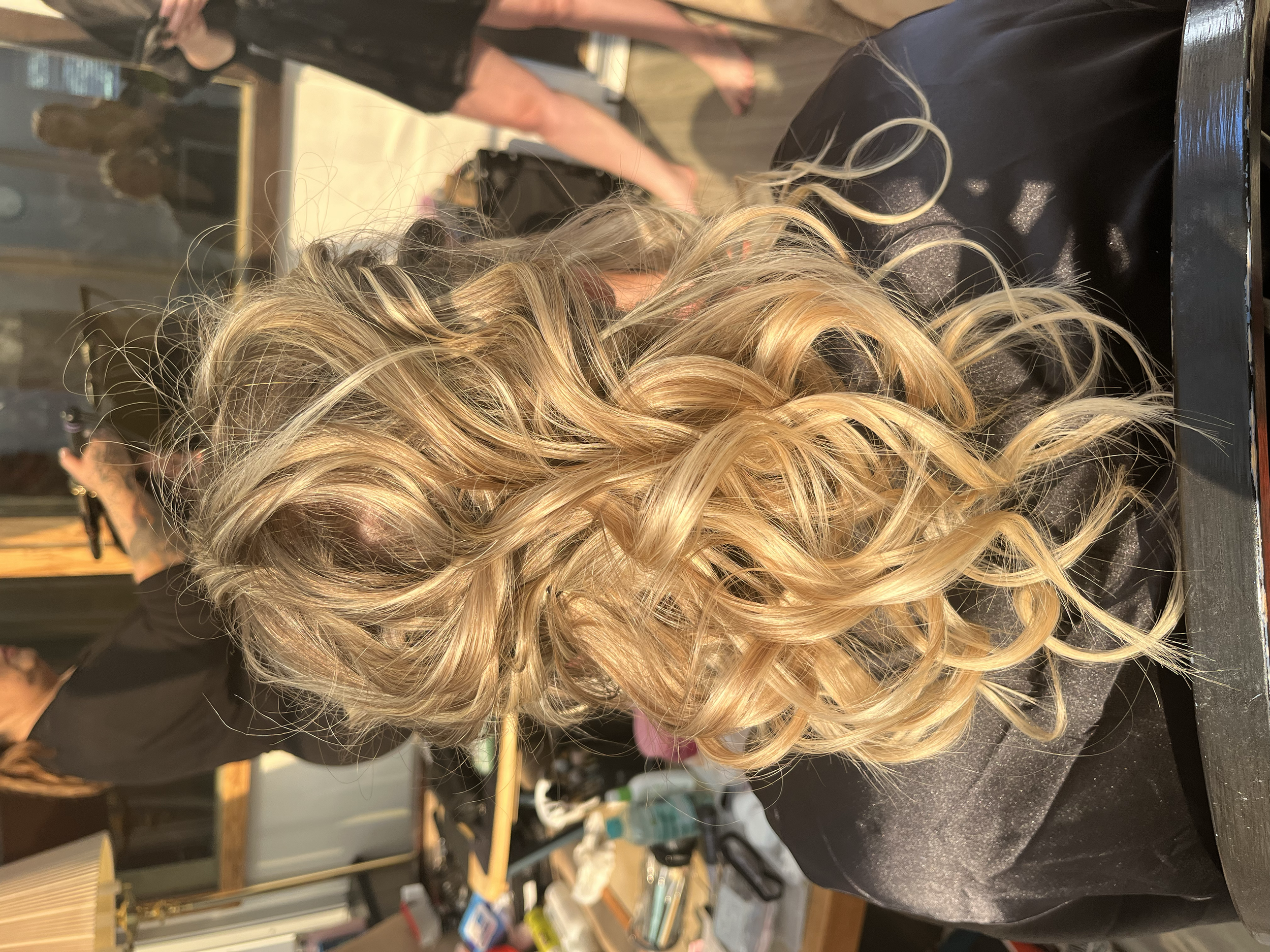 Person with long, blonde, curly hair sitting at a salon or hair studio, with hair styled in loose curls.