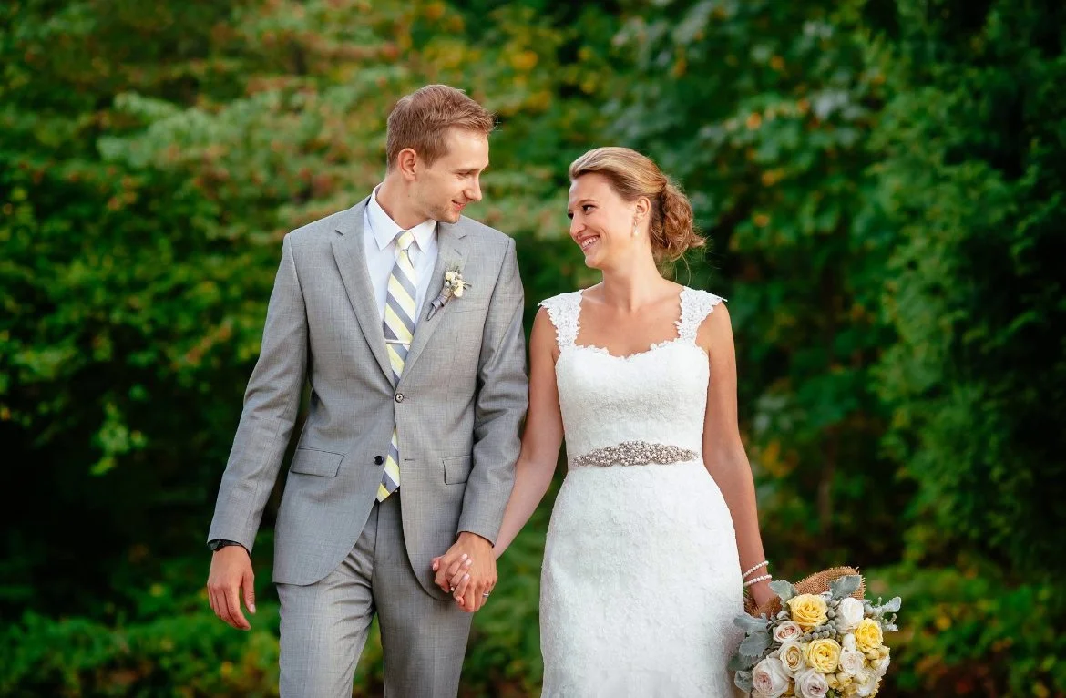 A bride and groom holding hands and smiling at each other outdoors with green foliage in the background.