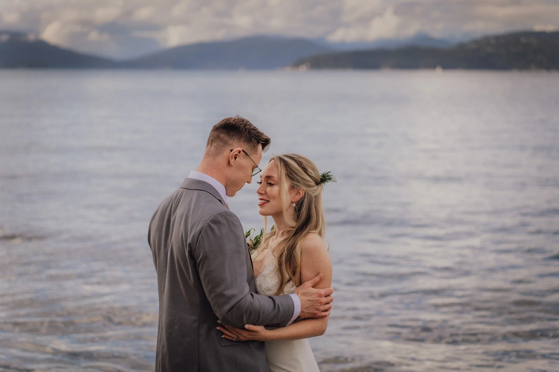 A newly married couple standing on a beach by the water, gazing at each other with their foreheads touching, during sunset or dusk.