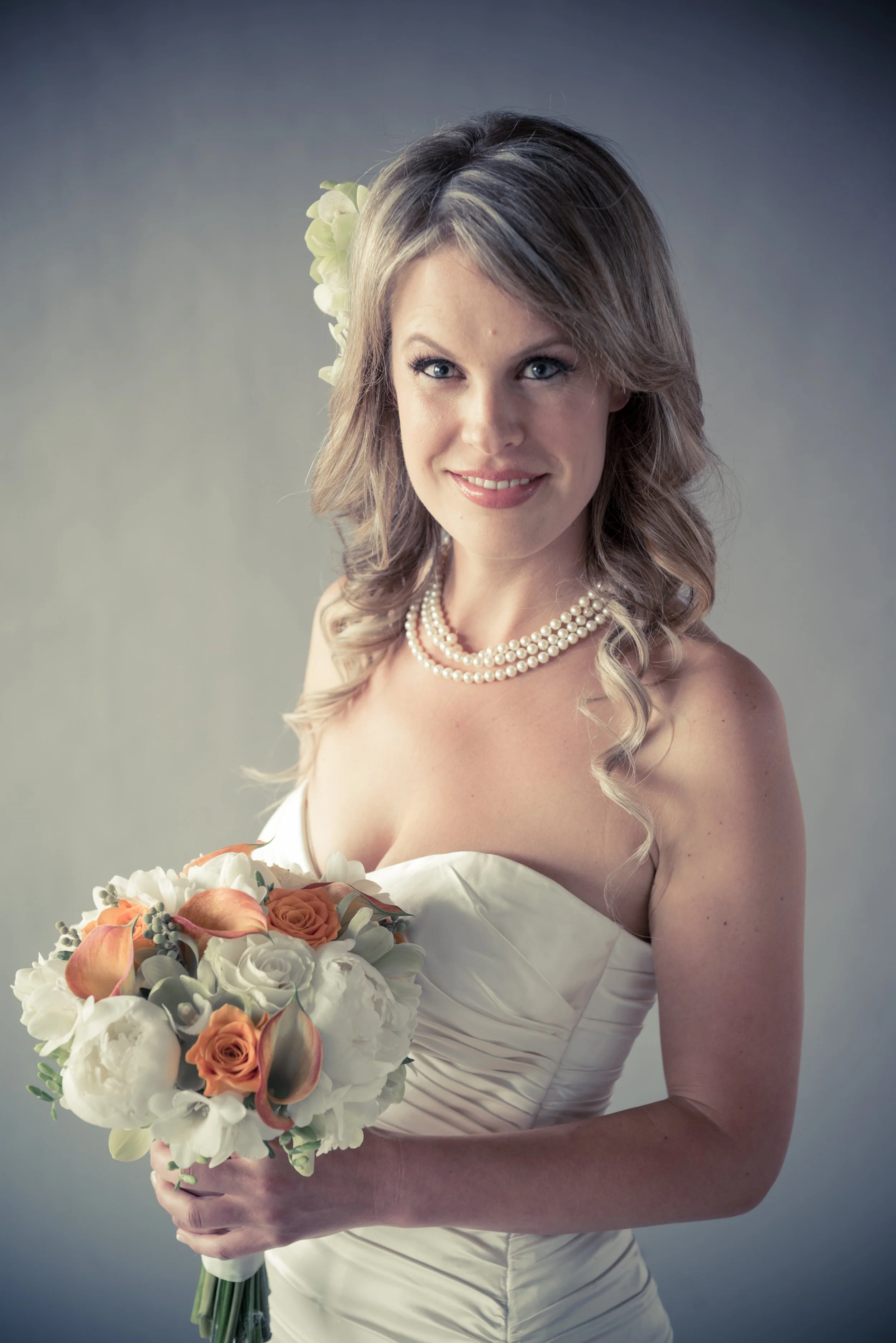 A woman in a strapless white wedding gown with pearl necklace holding a bouquet of white and orange flowers, with a blonde flower in her hair, smiling at the camera.