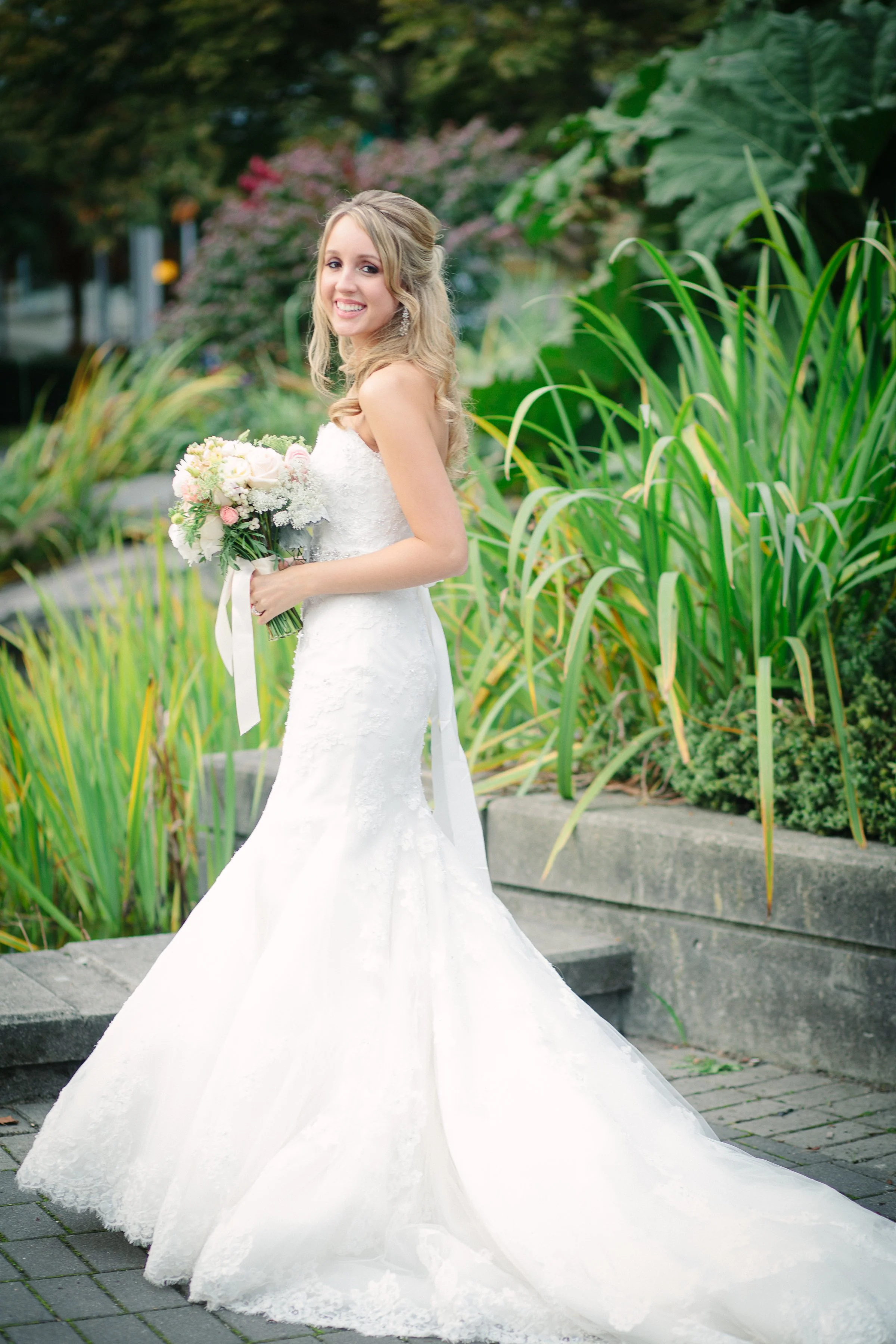 A smiling bride in a white lace wedding gown holding a bouquet stands outdoors among green plants and flowers.