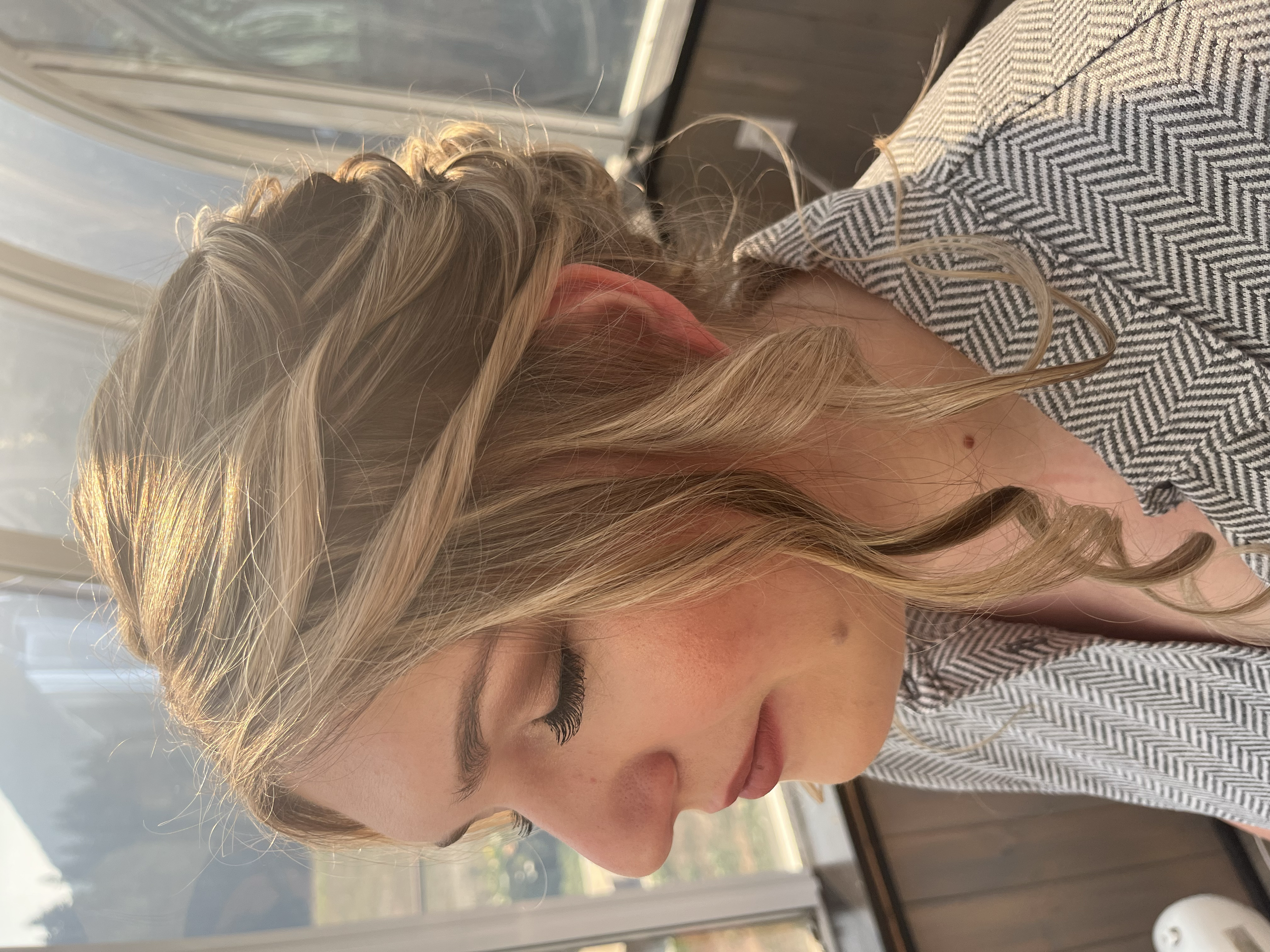 Close-up of a woman with light brown, wavy hair, wearing a striped shirt with a v-neck, sitting indoors near a window with a wooden wall behind her.