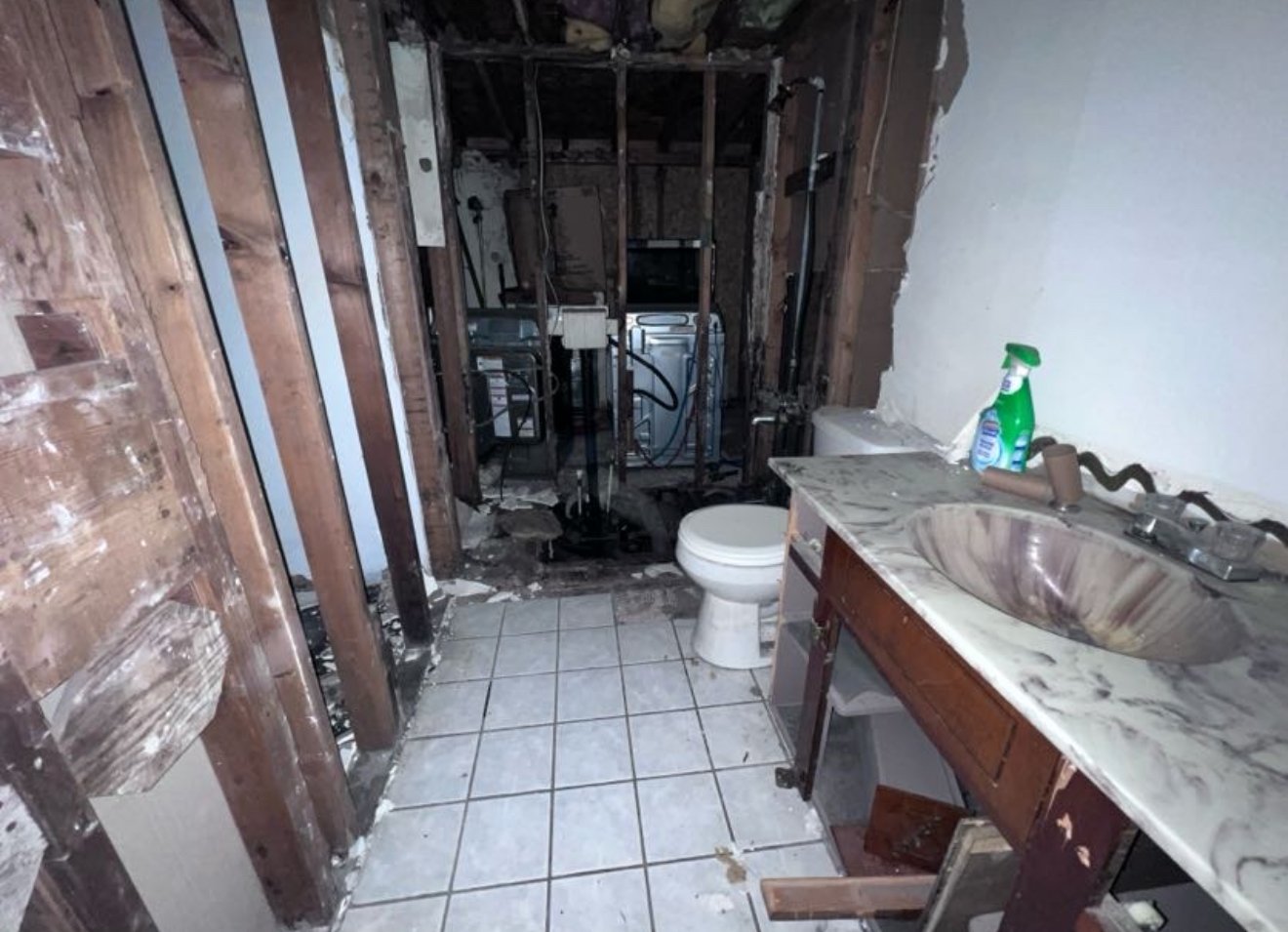 Bathroom in disrepair with damaged walls, missing fixtures, and burned area behind toilet. Sink countertop covered in debris, spray bottle, and cleaning supplies on the right side.