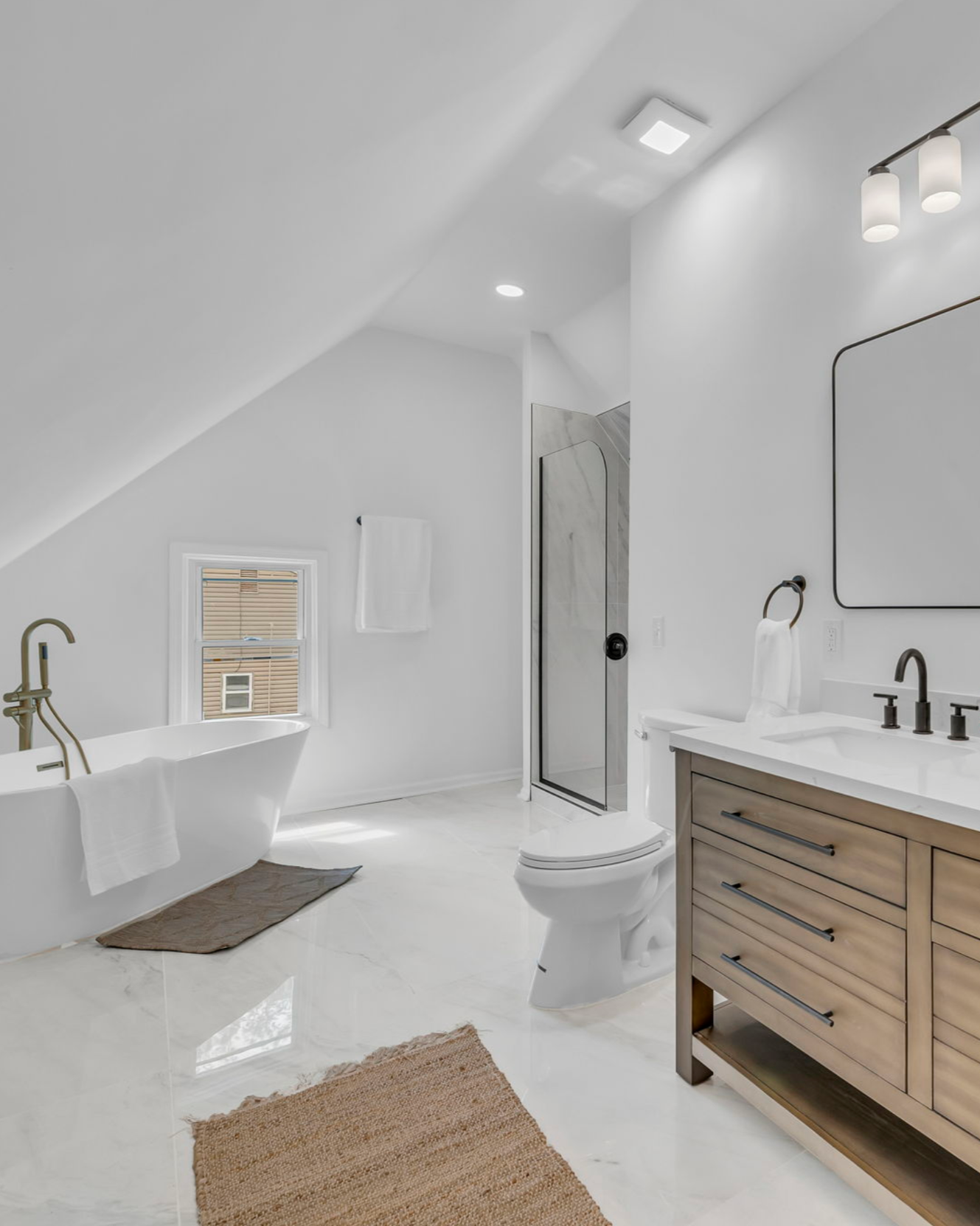 Modern white bathroom with a bathtub and brass faucet, a glass shower, a wooden vanity with black hardware, a large mirror, a toilet, a small window, and beige and brown rugs.