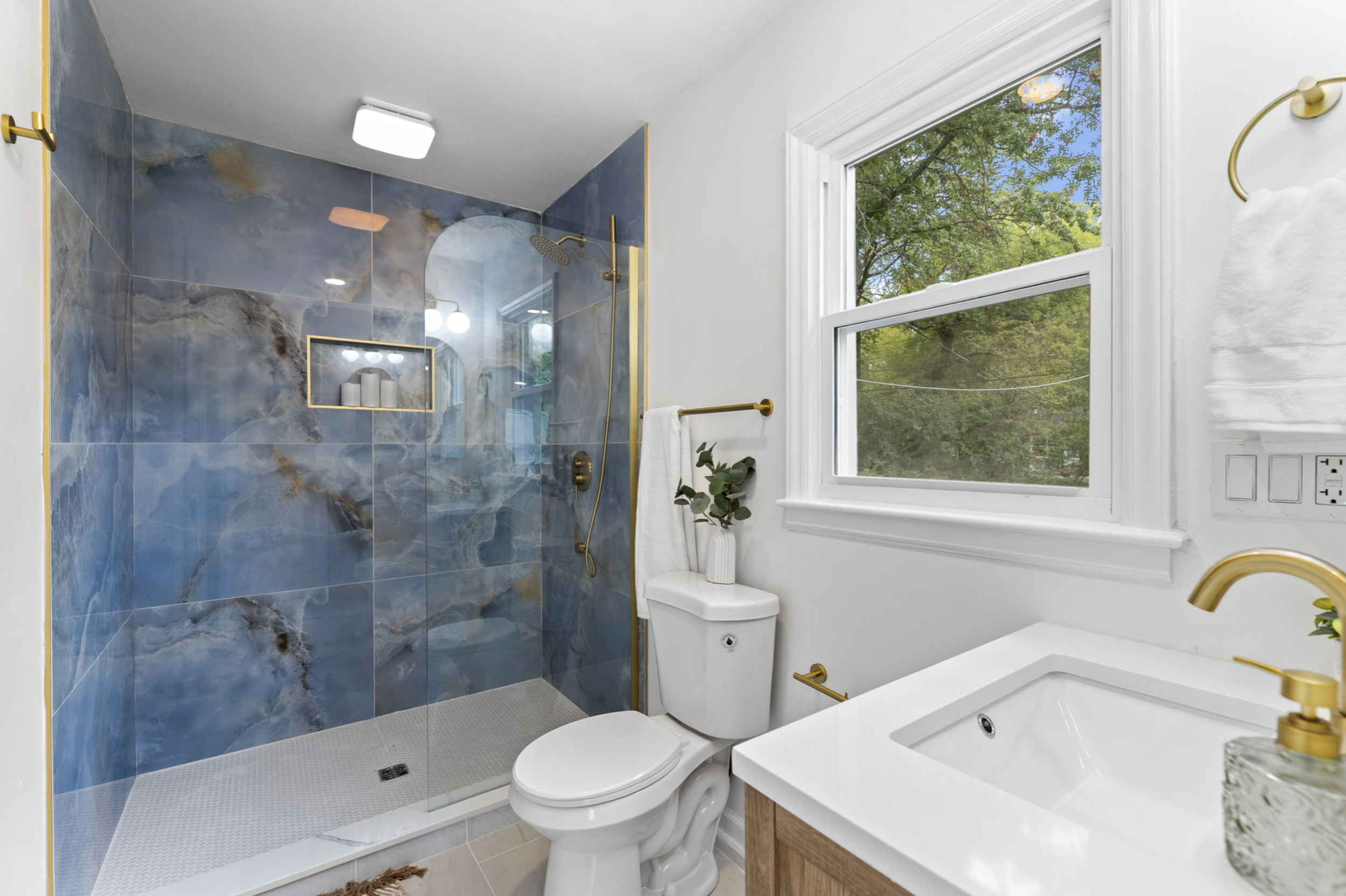 Modern bathroom with a walk-in shower featuring blue and gold marble tiles, a white toilet, a wooden vanity, and a large window showing green trees outside.