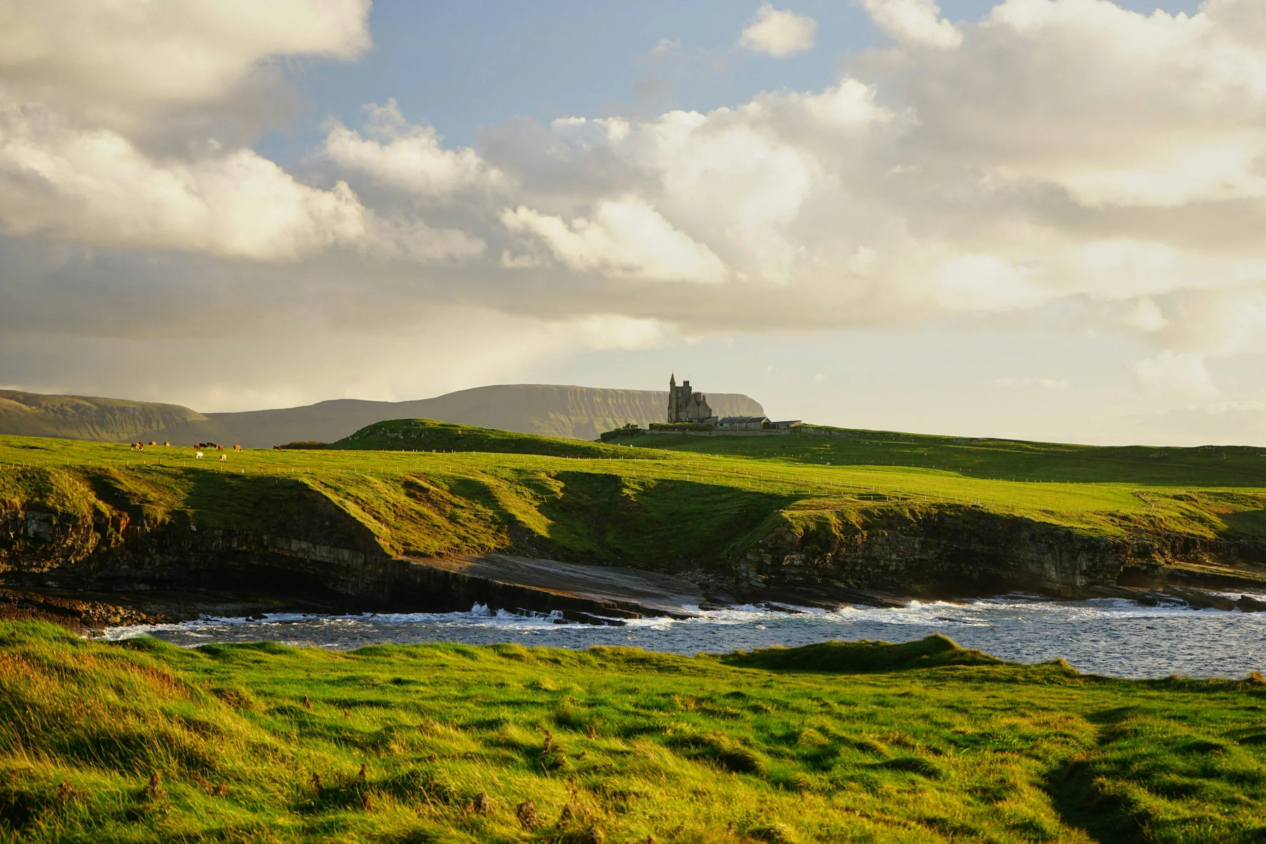 A scenic landscape of green grassy fields with a castle on a distant hill, behind cliffs overlooking the ocean under a partly cloudy sky.