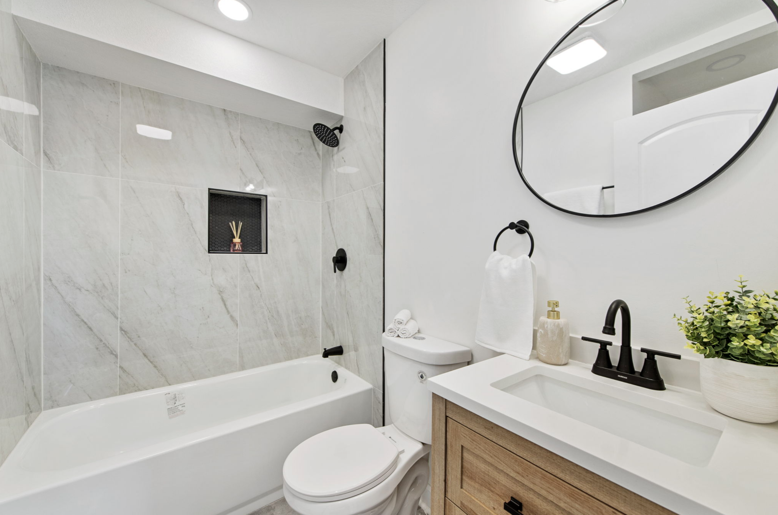 Modern bathroom with white and beige color scheme, black fixtures, oval mirror, potted plant, and minimalist decor.