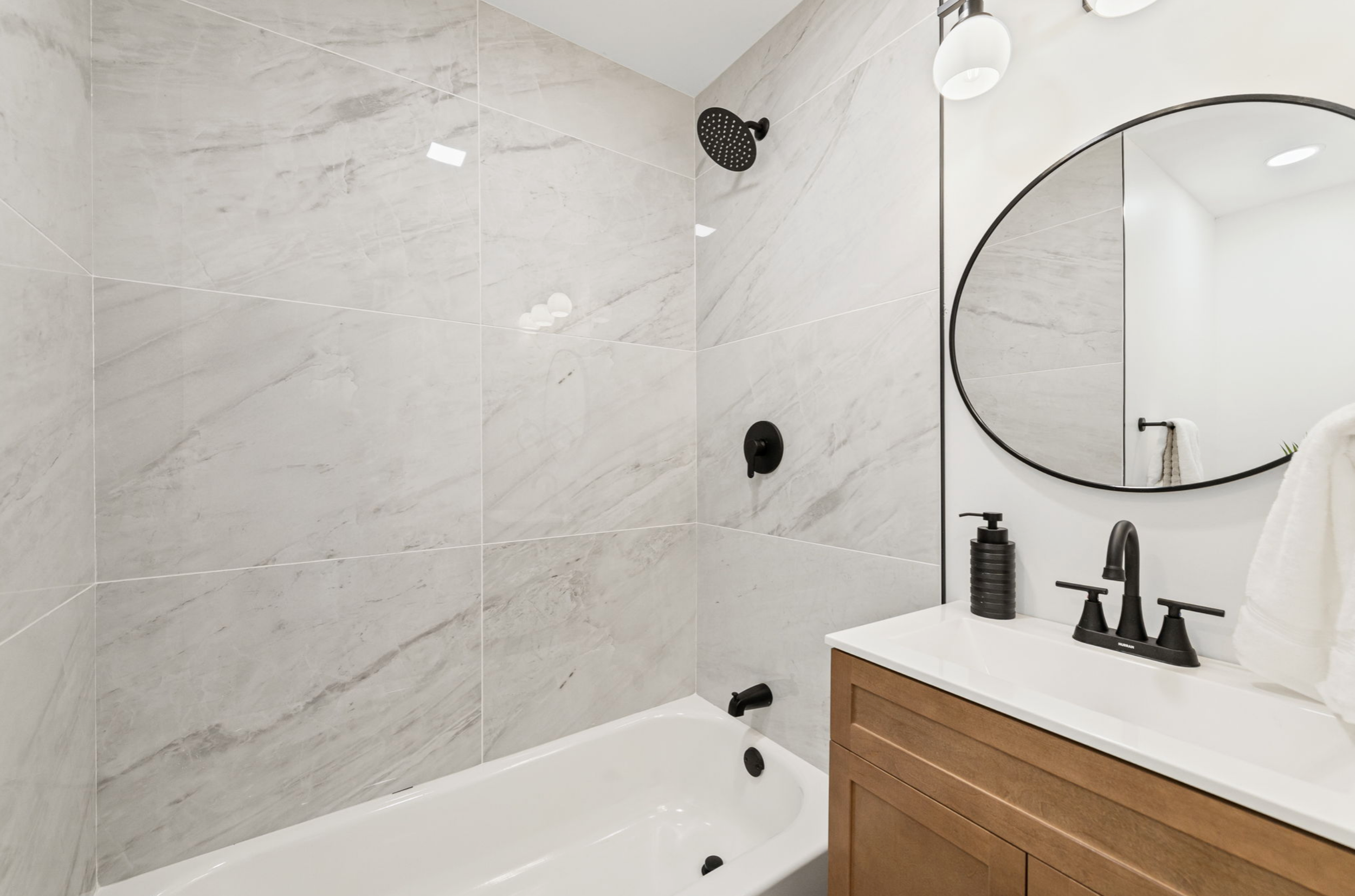Modern bathroom with a white bathtub, marble tiled wall, black showerhead, and a wooden vanity with a white sink. A round mirror is mounted above the sink, and a black soap dispenser sits on the countertop.