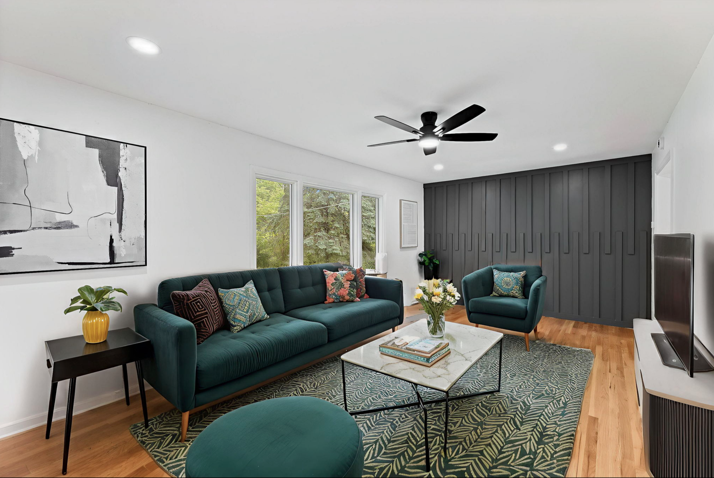 Living room with green sofa and armchair, black side table with yellow pot, white marble coffee table, abstract black-and-white artwork, three windows showing trees outside, dark gray accent wall, ceiling fan, flat-screen TV, wooden floor, and decora