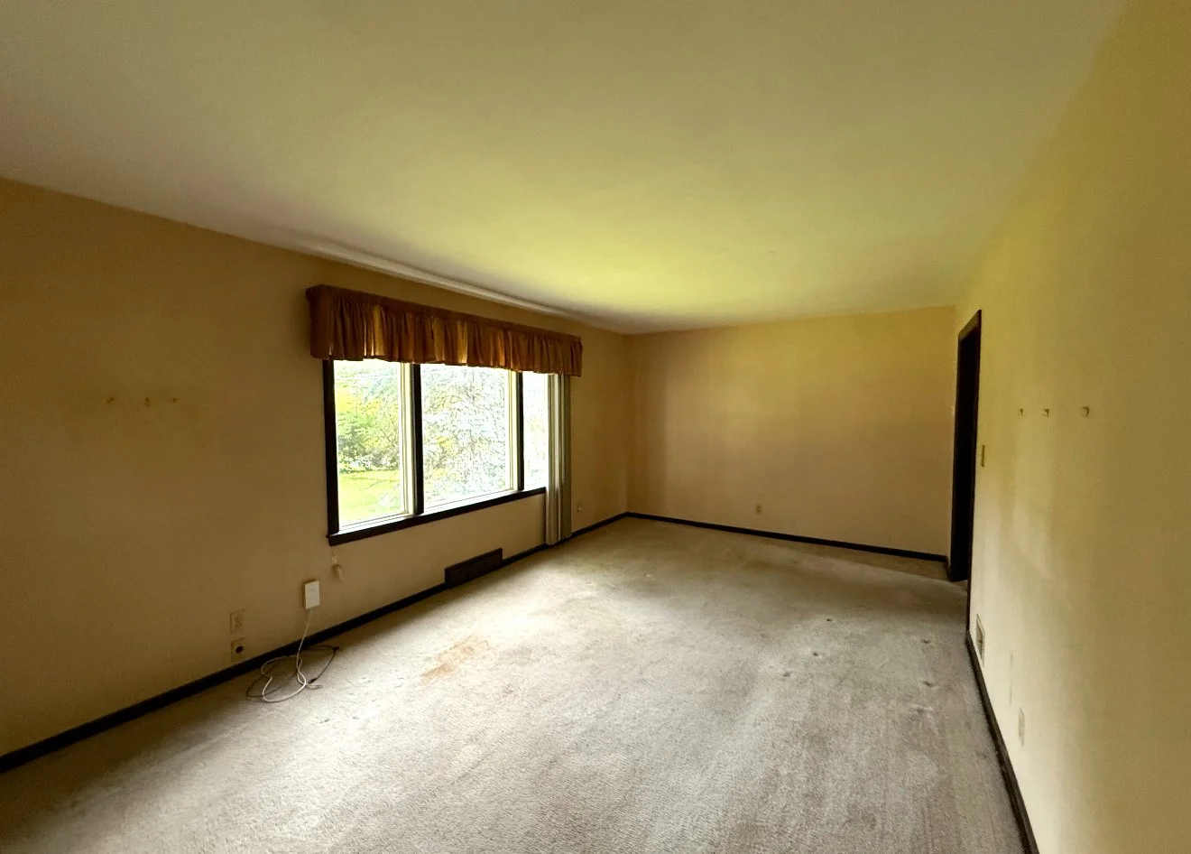Empty living room with beige carpet, yellow walls, large window with a brown valance, and a doorway on the right.