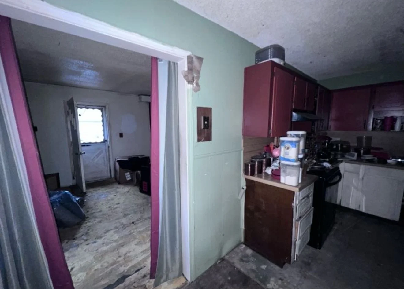 Interior view of a cluttered kitchen and an adjoining room, with visible cabinets, countertop appliances, and a doorway leading to a small room with a window.