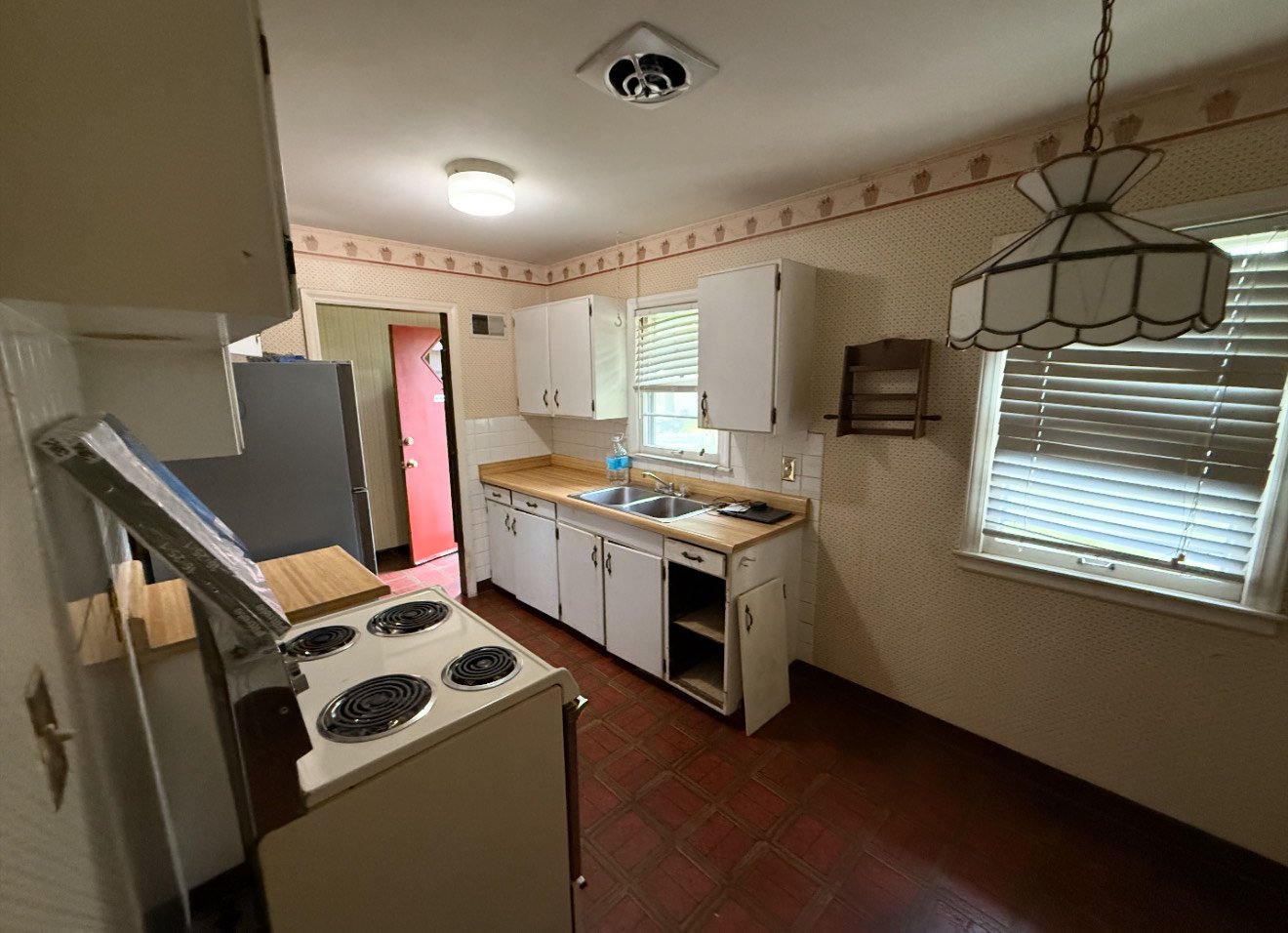 Older kitchen with white cabinets, a double sink, wooden countertop, window with blinds, an electric stove, and vintage light fixtures.