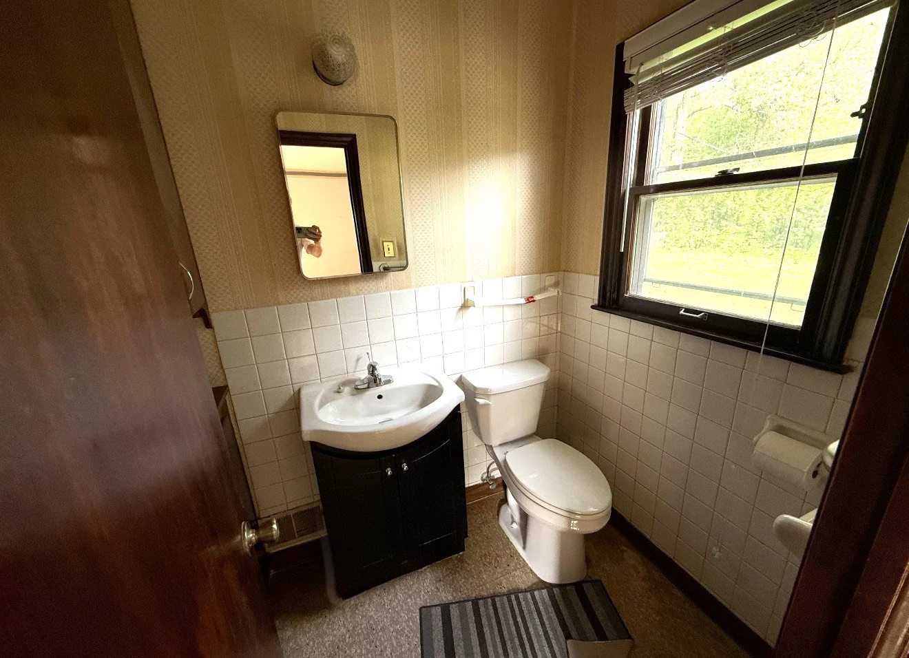 A small bathroom with a window showing trees outside, a white toilet, a small white sink with a black cabinet, a mirror above the sink, a bath mat on the floor, and a partial view of a wooden door.