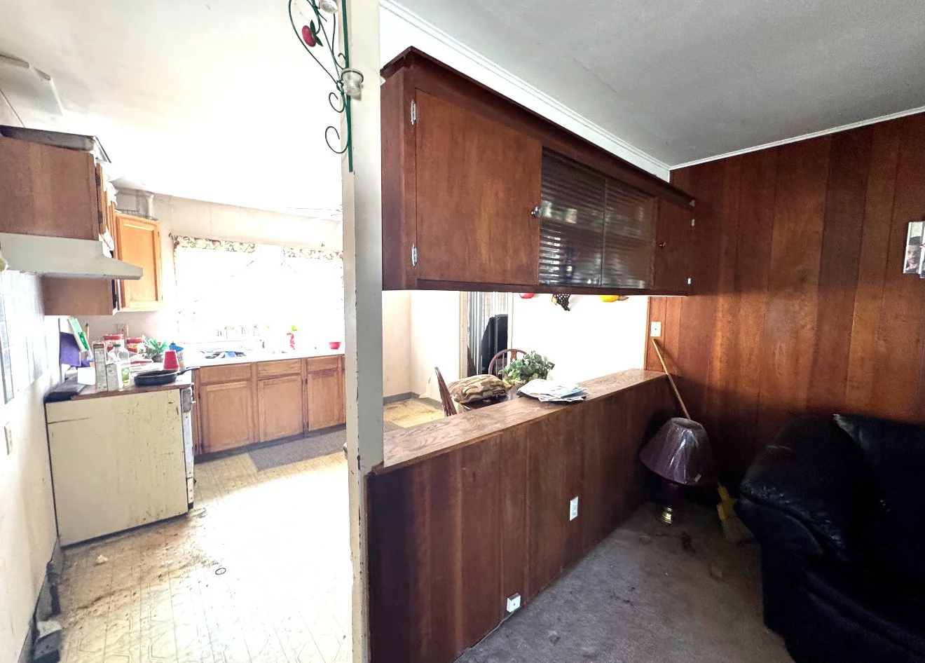 View of a kitchen and living room in a modest home. The kitchen has wooden cabinets, a white countertop with assorted items, and a window with a floral curtain. The living room features wood-paneled walls, a black couch, a lamp, and a small wooden ta