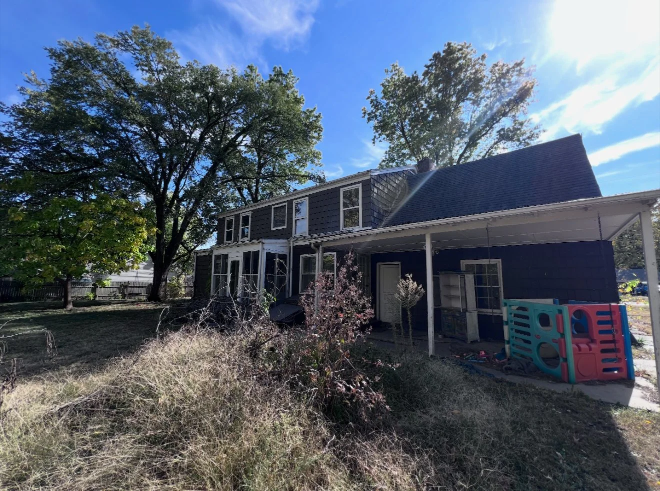 Backyard view of a two-story house with a covered patio, surrounded by trees and dry grass, sunny with a blue sky.