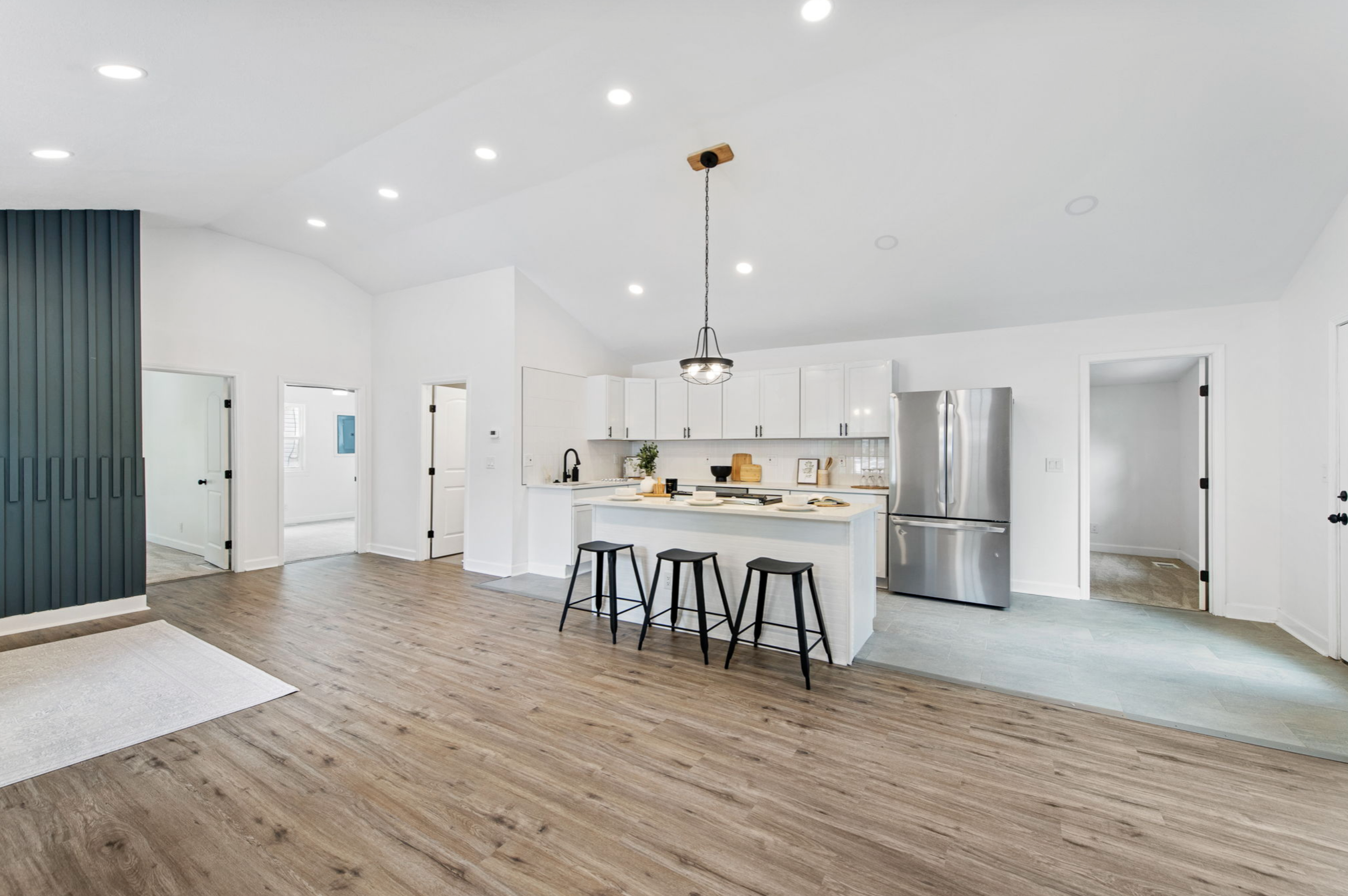 Open-plan kitchen with white cabinets, stainless steel refrigerator, black countertop faucet, island with three black barstools, and wood flooring.